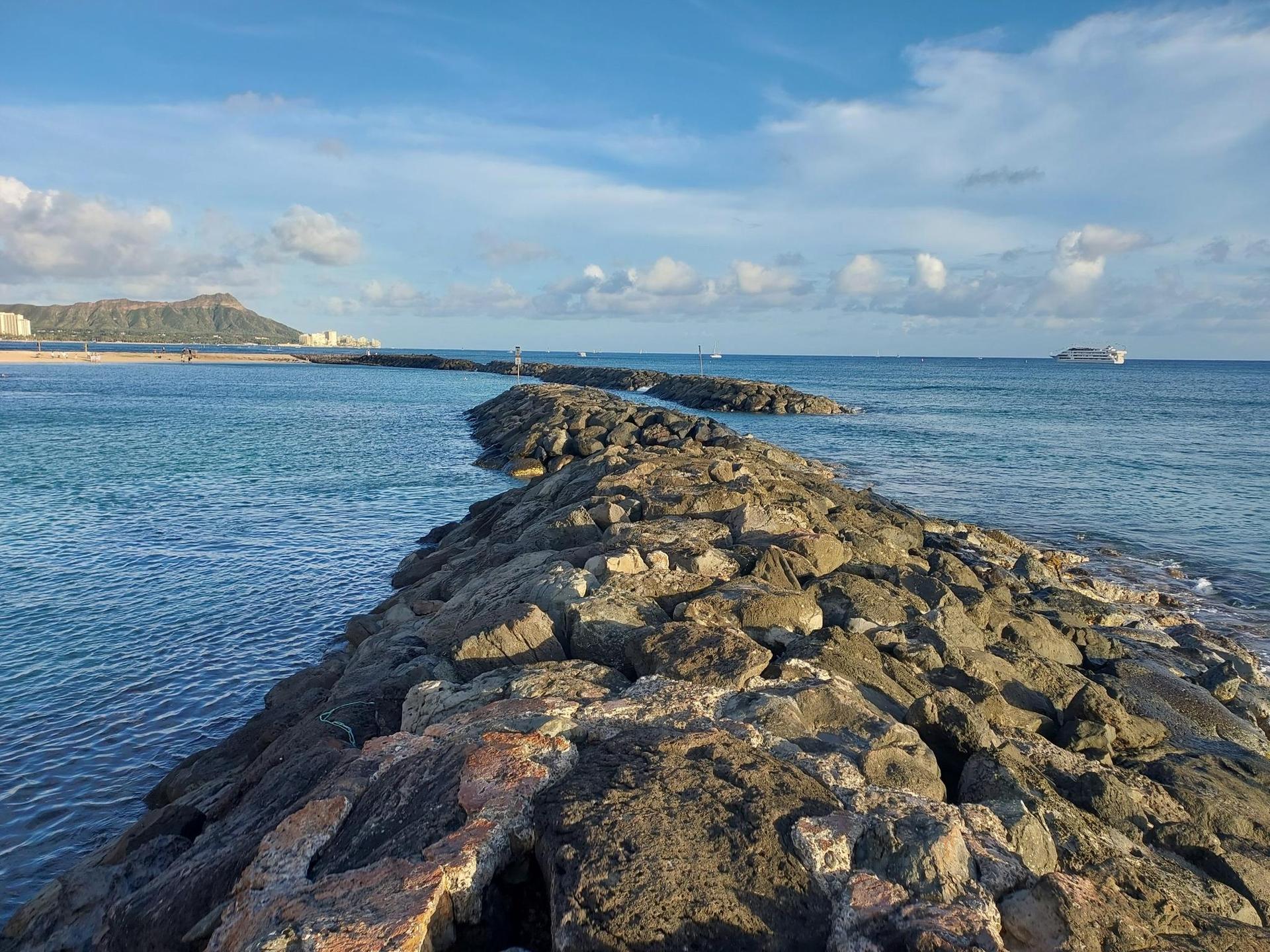 Rocky breakwater extends into bright blue ocean under a partly cloudy sky.