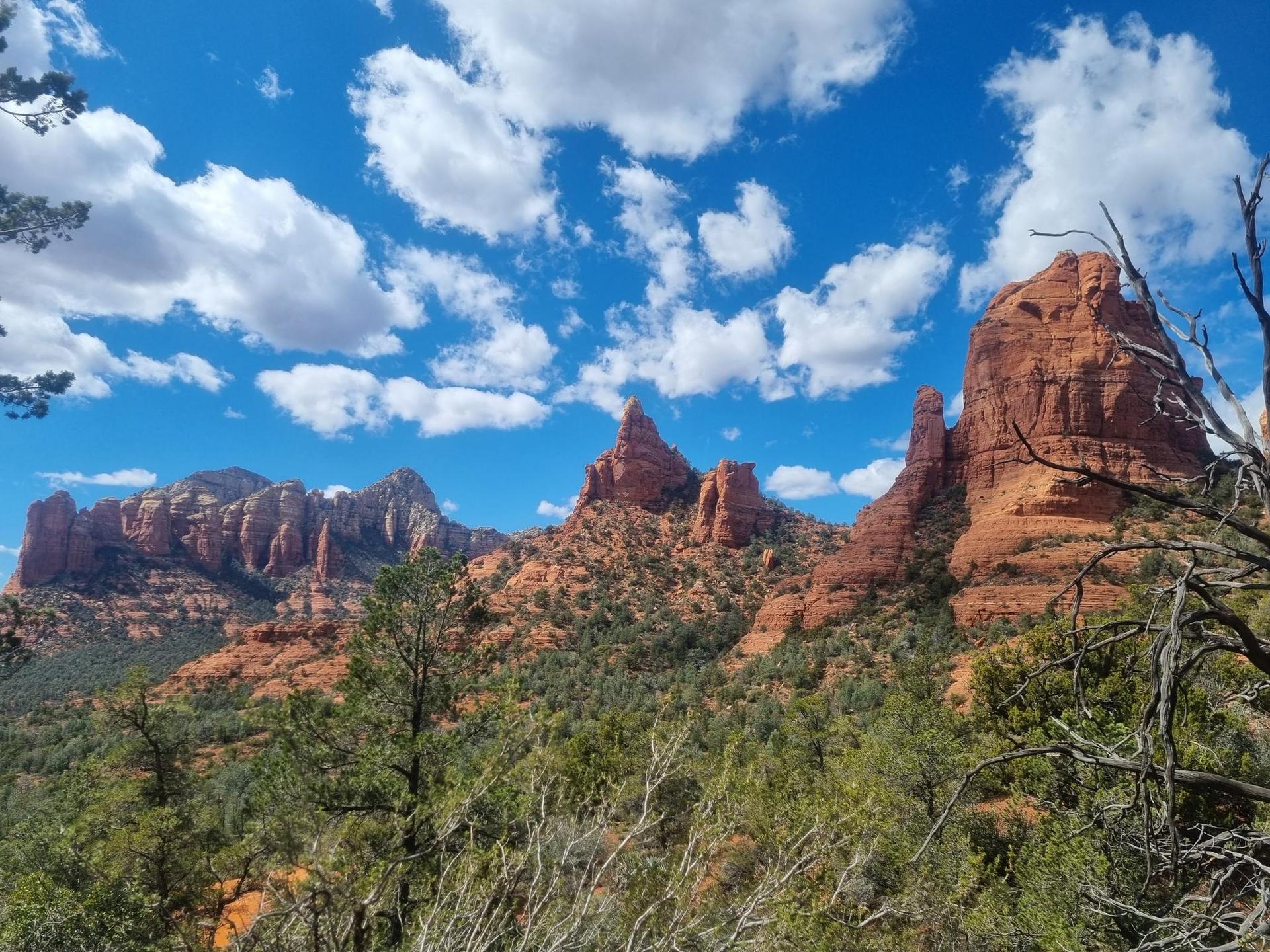 Majestic red rock formations and green trees under a bright blue, cloudy sky.