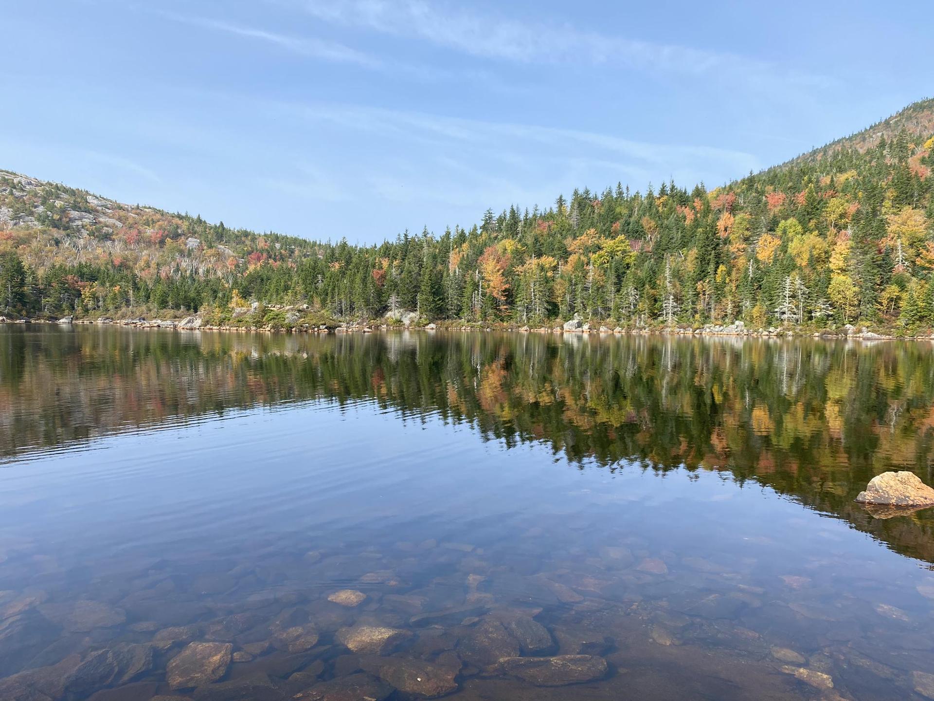 Calm lake reflecting autumn trees and mountains under a clear blue sky.