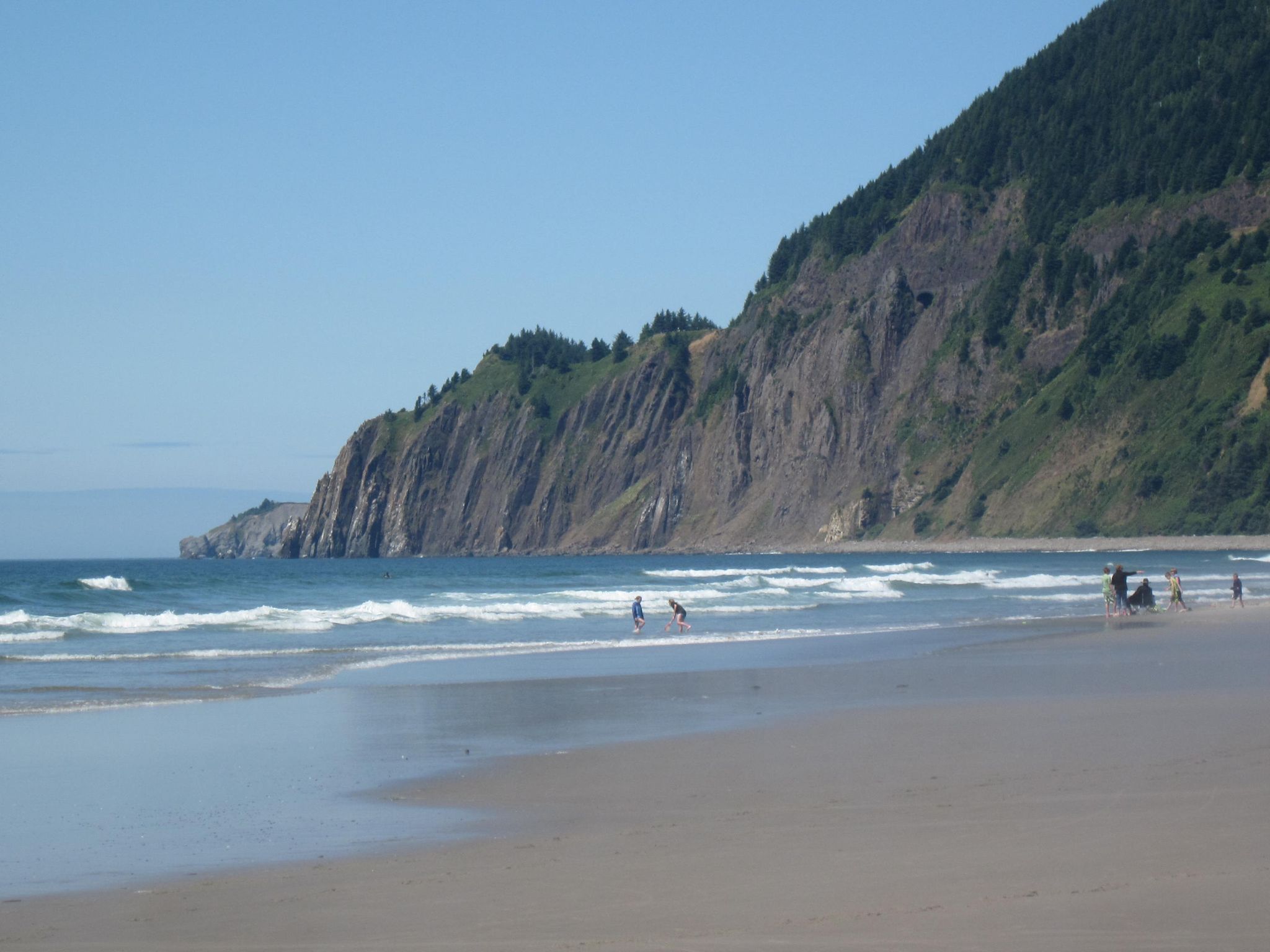 Sunny beach with a large forested cliff and people walking near the waves. - Manzanita Beach loop from Manzanita