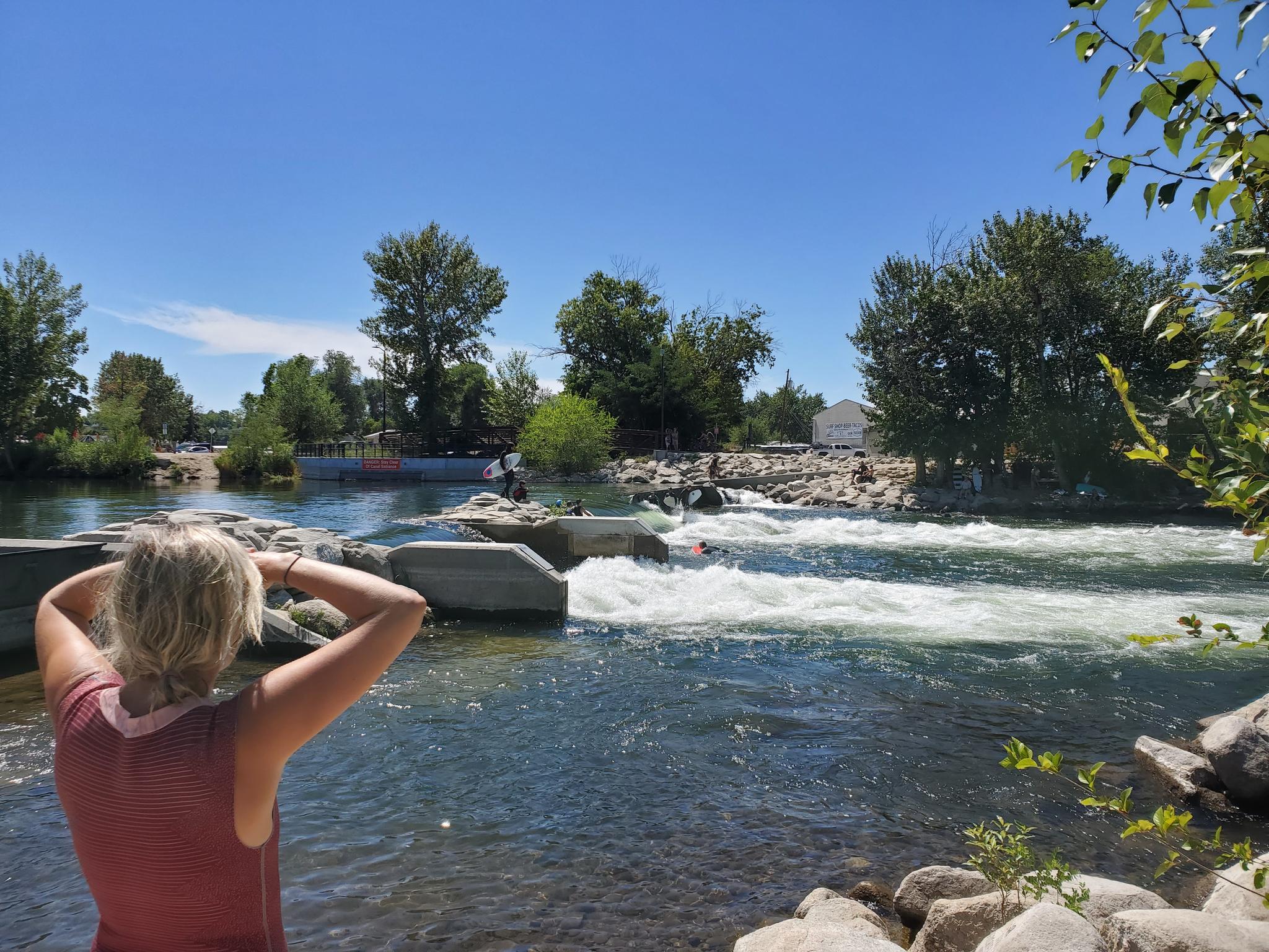 Woman views a sunny river with rapids and distant people on the water.