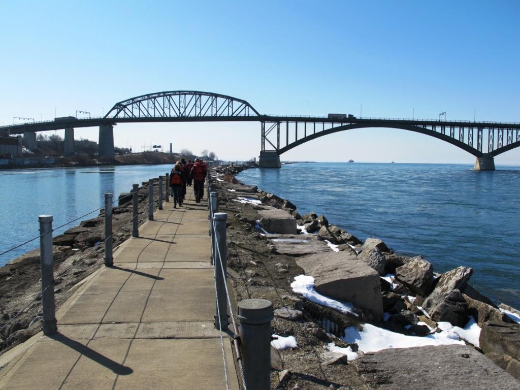 People walk on a concrete pier towards a large arched bridge over blue water.