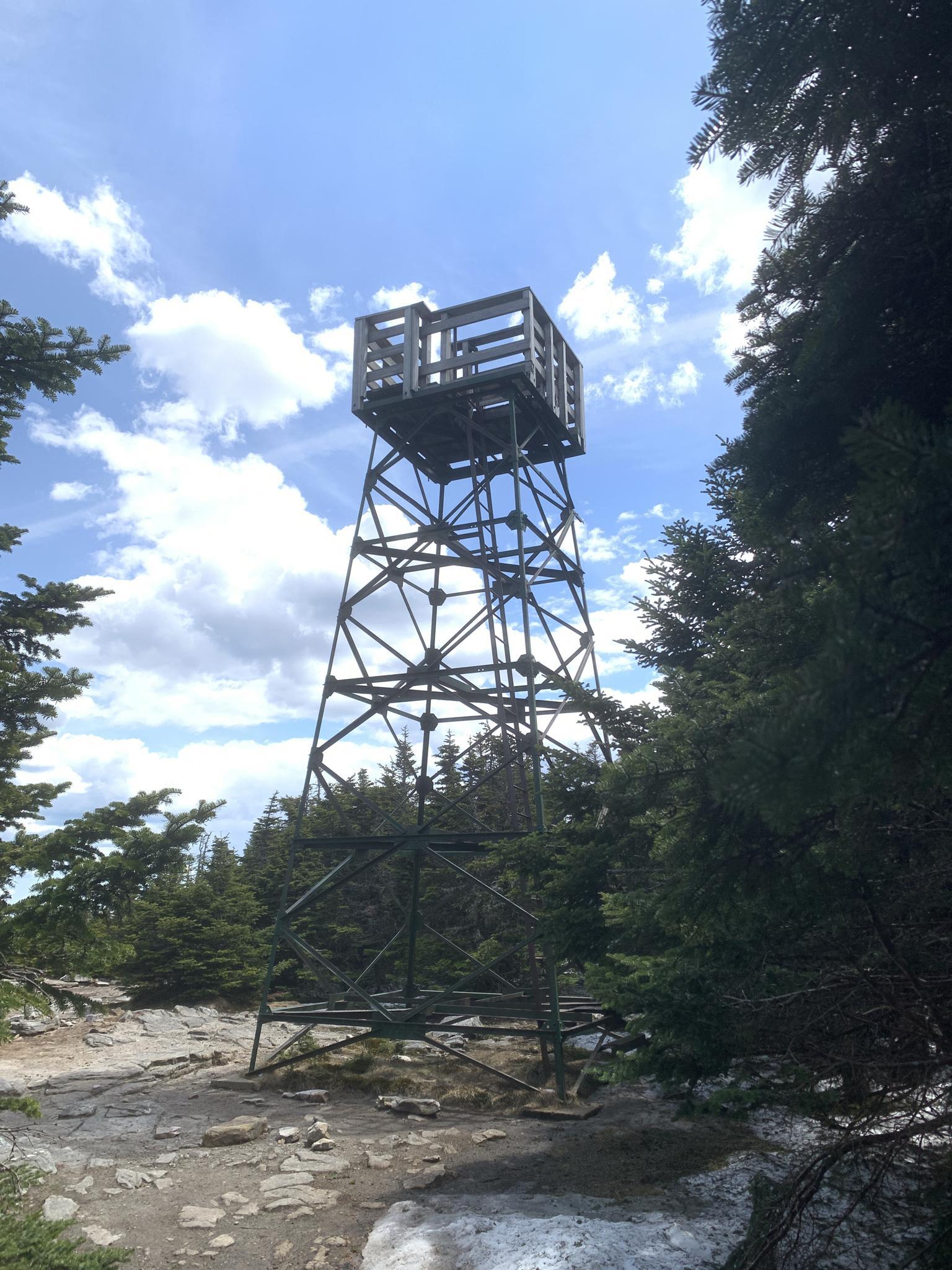 Tall metal fire tower on a rocky summit surrounded by evergreen trees.