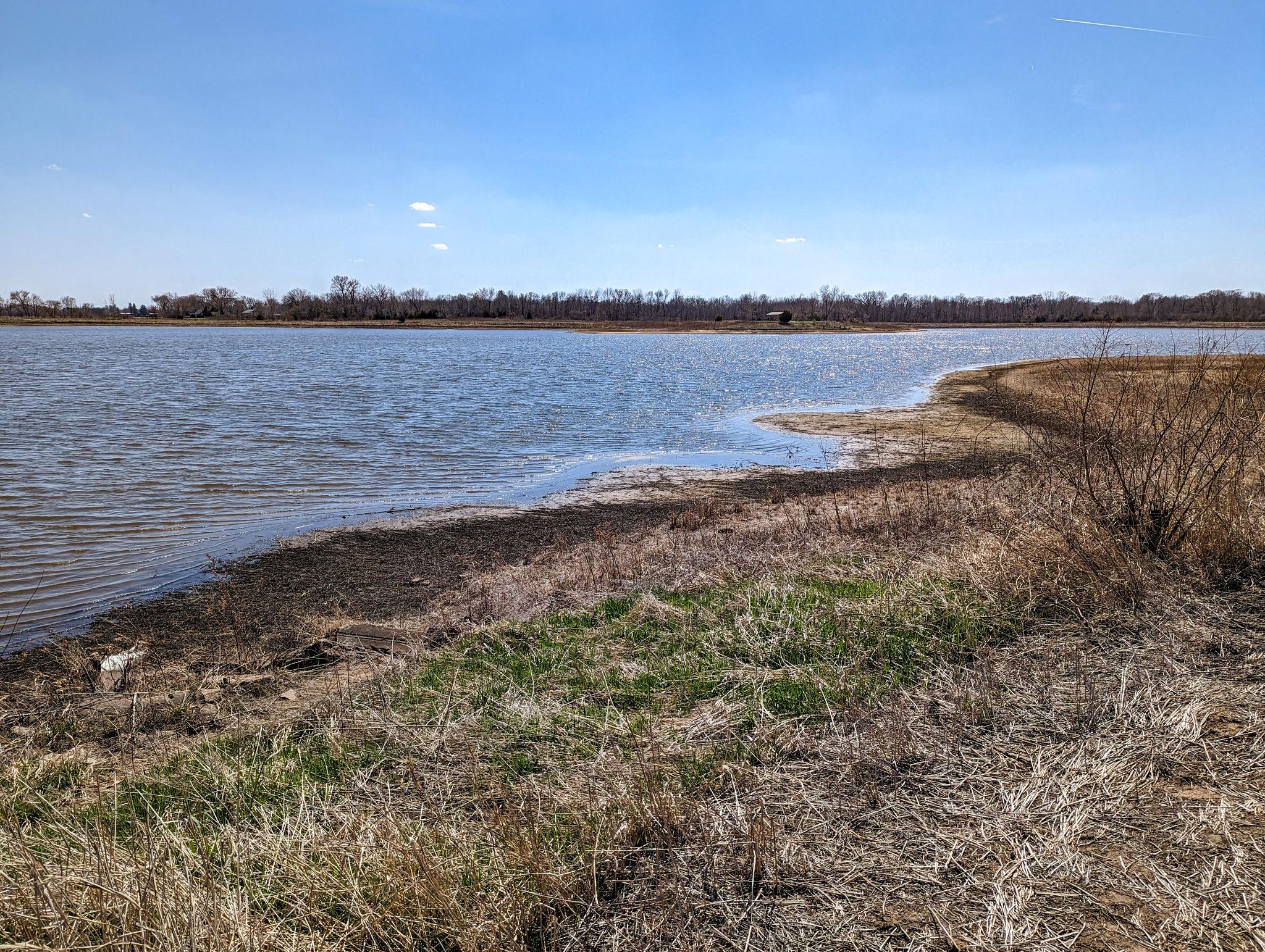 Wide lake with a dry grassy shore and distant bare trees under a clear blue sky.