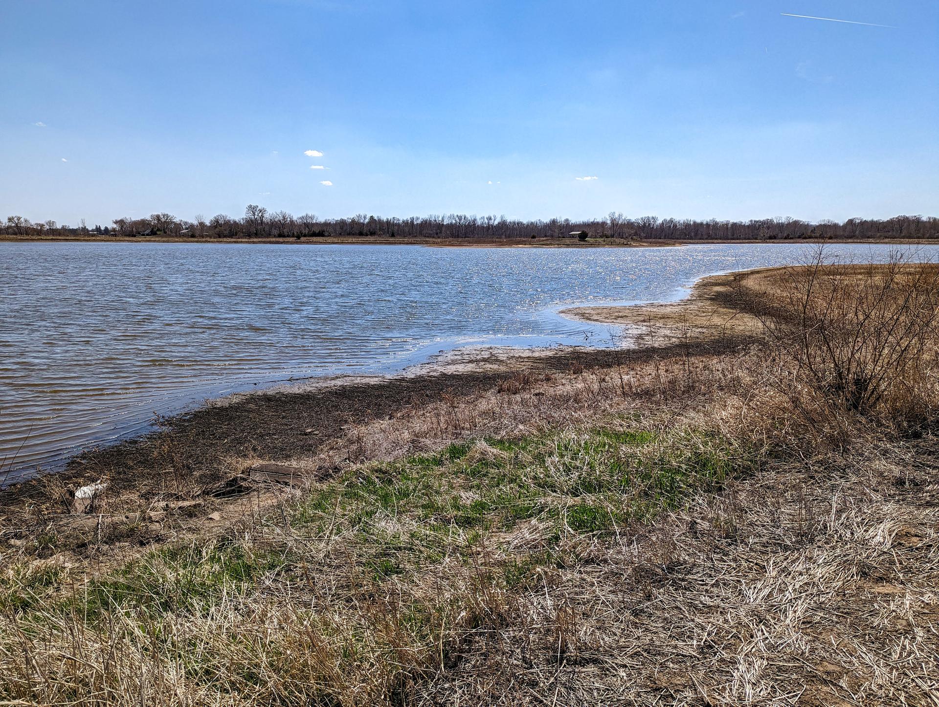 Wide lake with a dry grassy shore and distant bare trees under a clear blue sky.