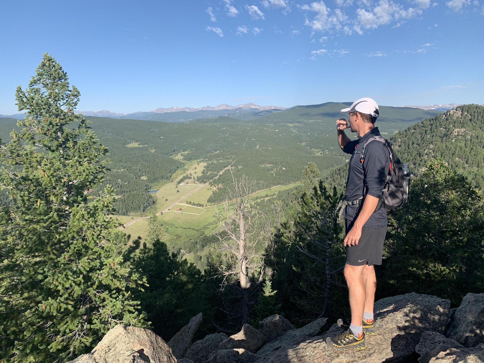 Man on a rocky outcrop views a vast, sunny, forested valley.
