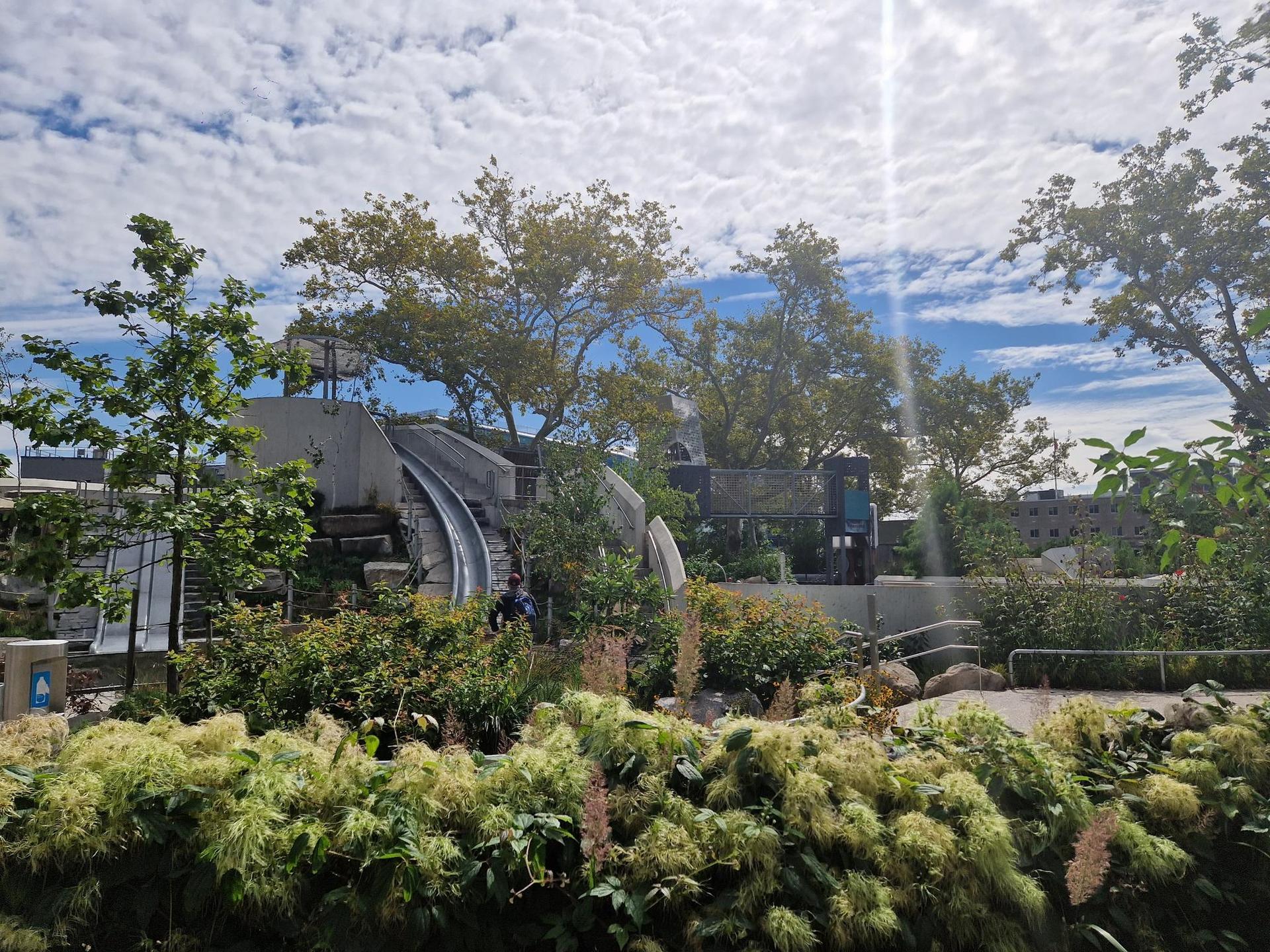 Sunny playground with slides and green bushes under a bright, cloudy sky.