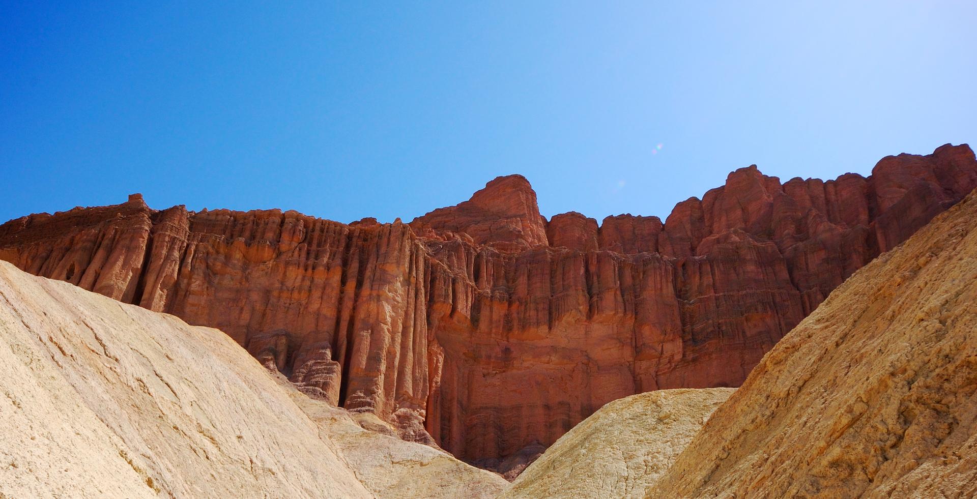 Steep, reddish-brown fluted cliffs rise above light-colored hills under a clear blue sky.