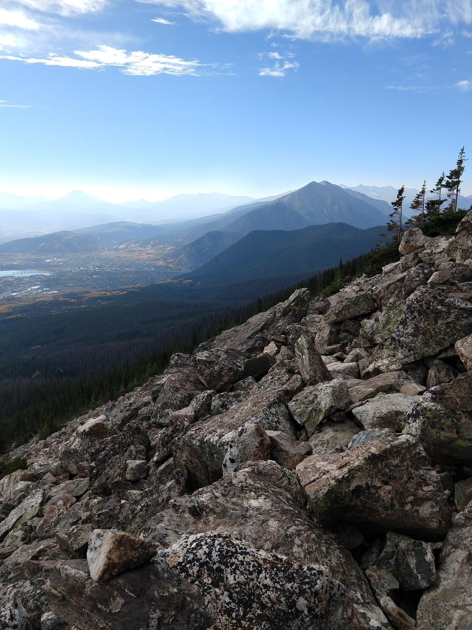 Rocky mountain slope with pine trees overlooking a hazy valley and distant lake.