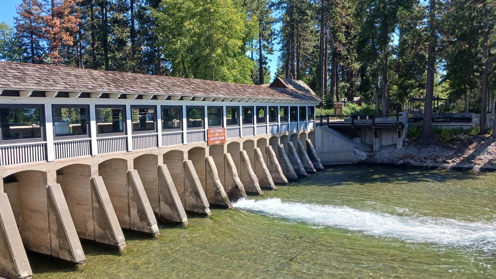 Covered dam structure with water flowing, surrounded by green pine trees.