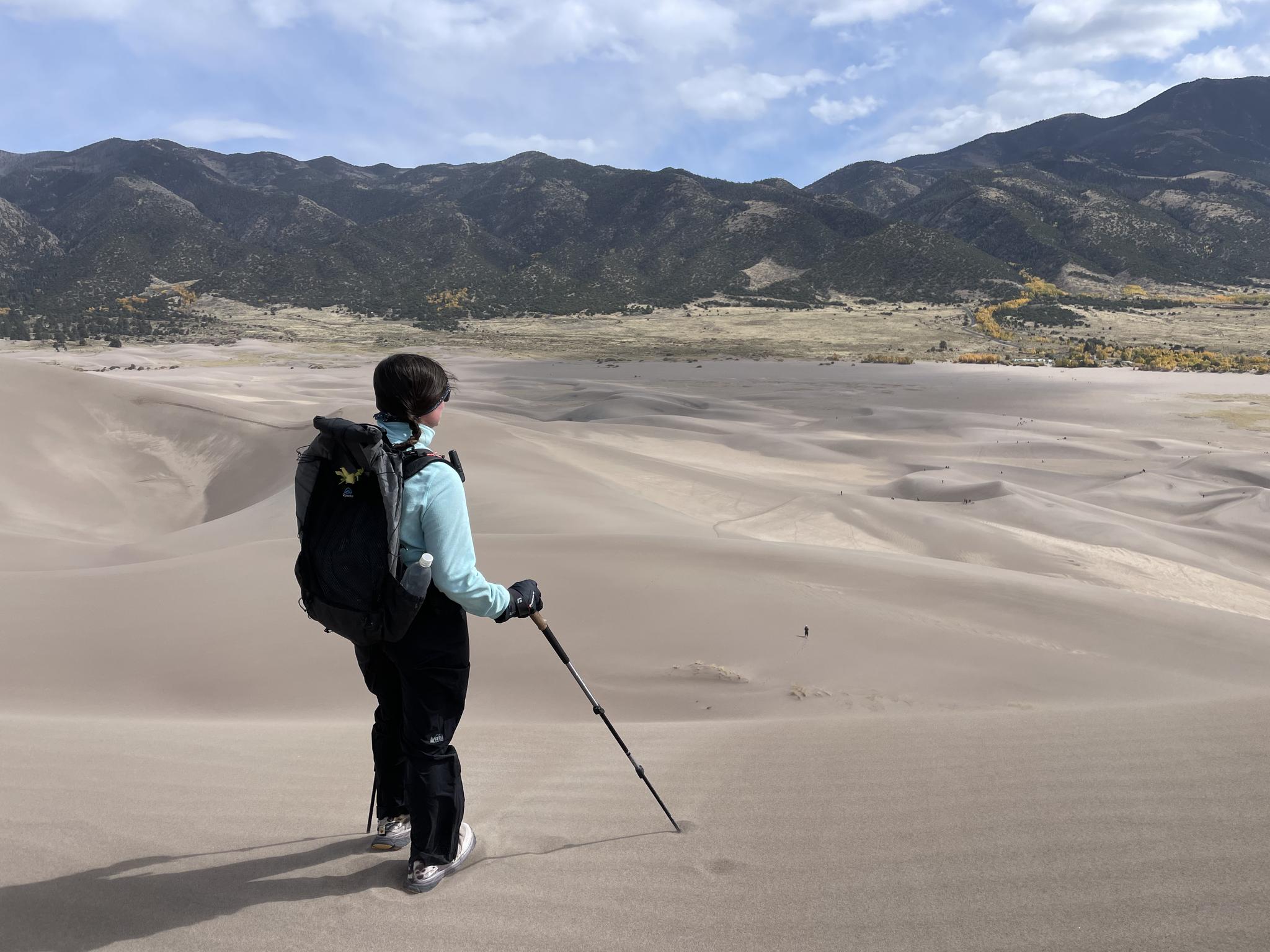 A hiker with a backpack and pole stands on a vast sand dune, mountains in distance.