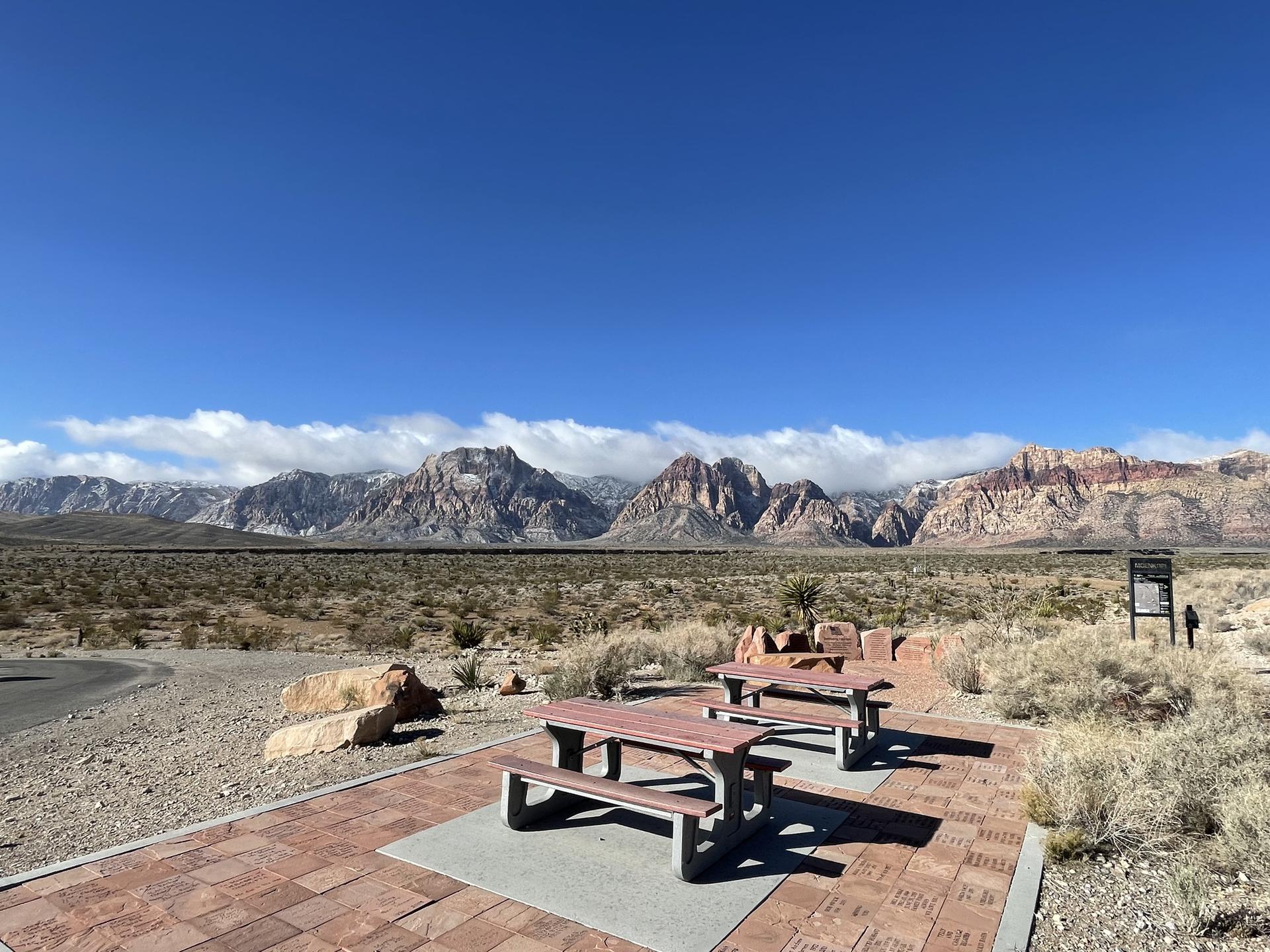Two picnic tables on a paved area, facing distant mountains under a clear blue sky.