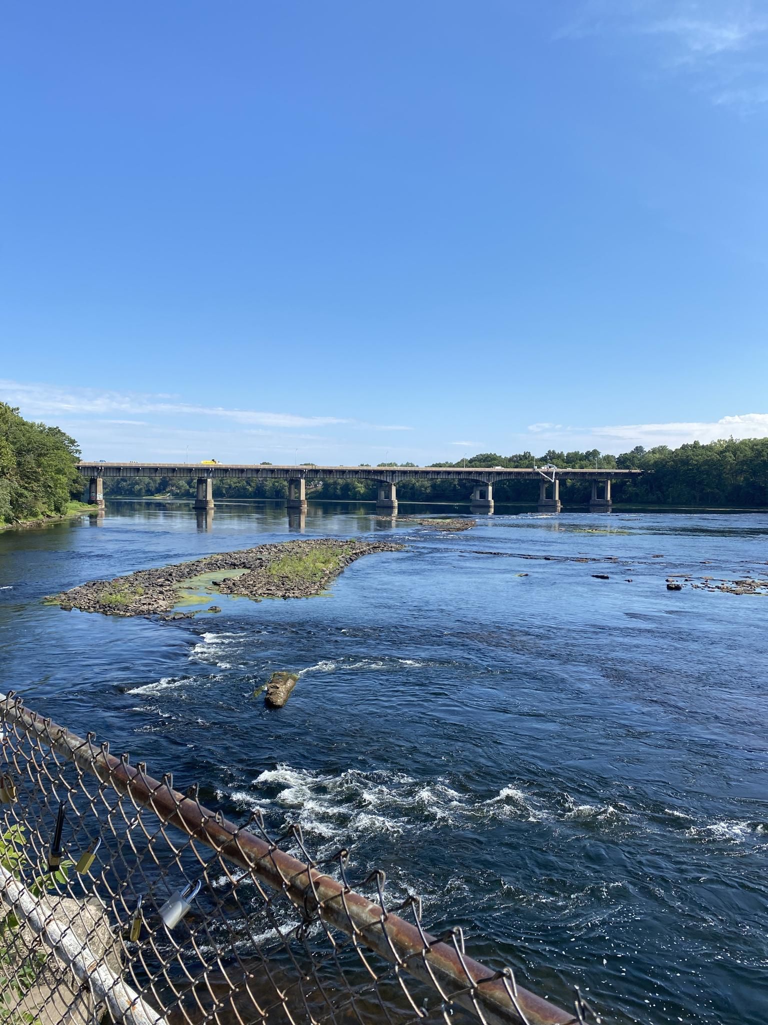 Bright daylight view of a river, distant bridge, and foreground fence. - Windsor Locks Canal State Park Trail