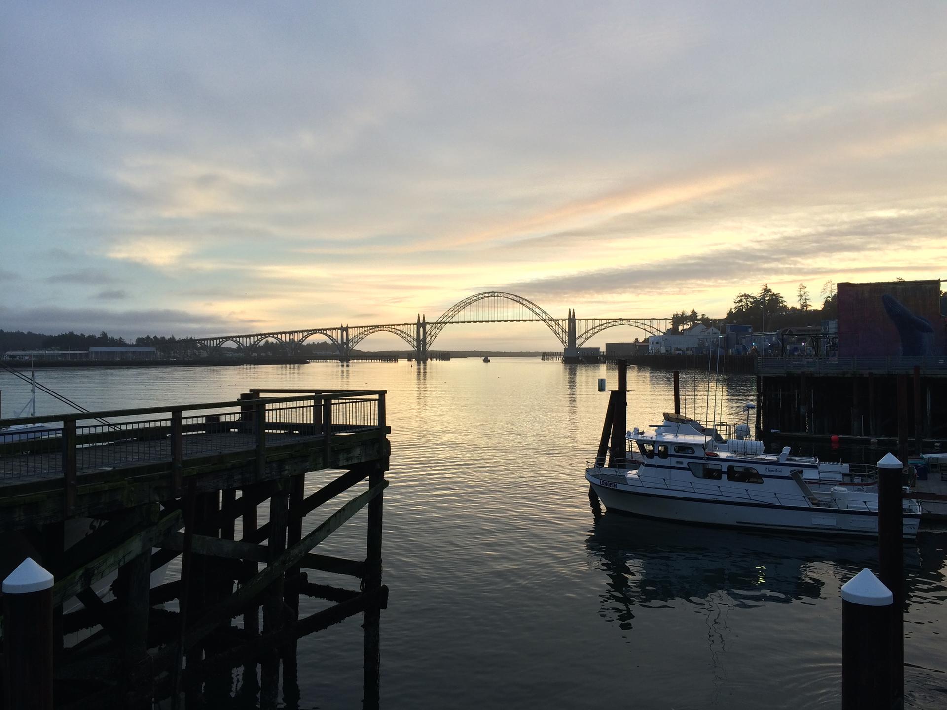 Arch bridge over calm water with boats docked at sunset or sunrise.