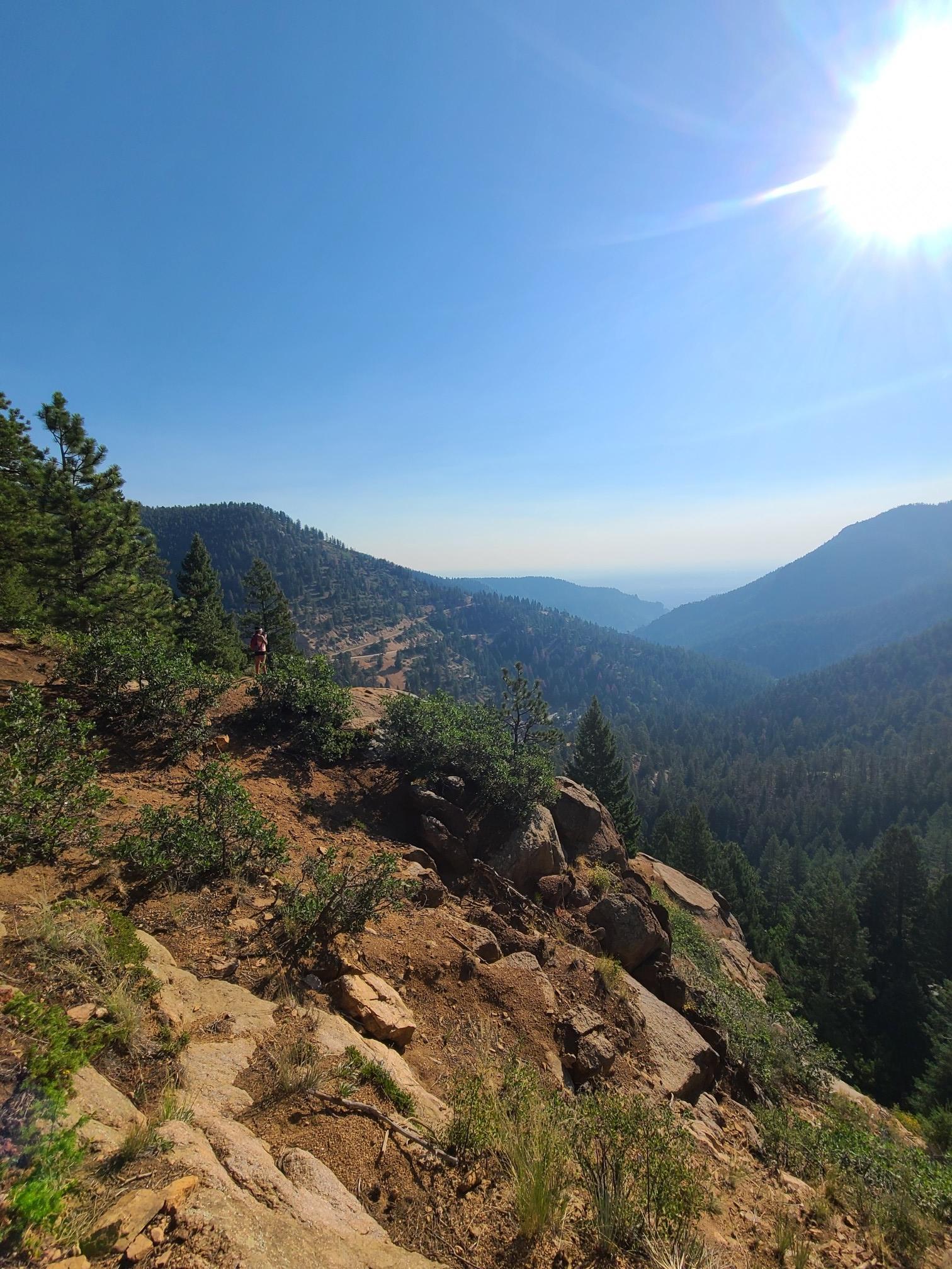 A person overlooks a vast forested valley and mountains on a sunny day.