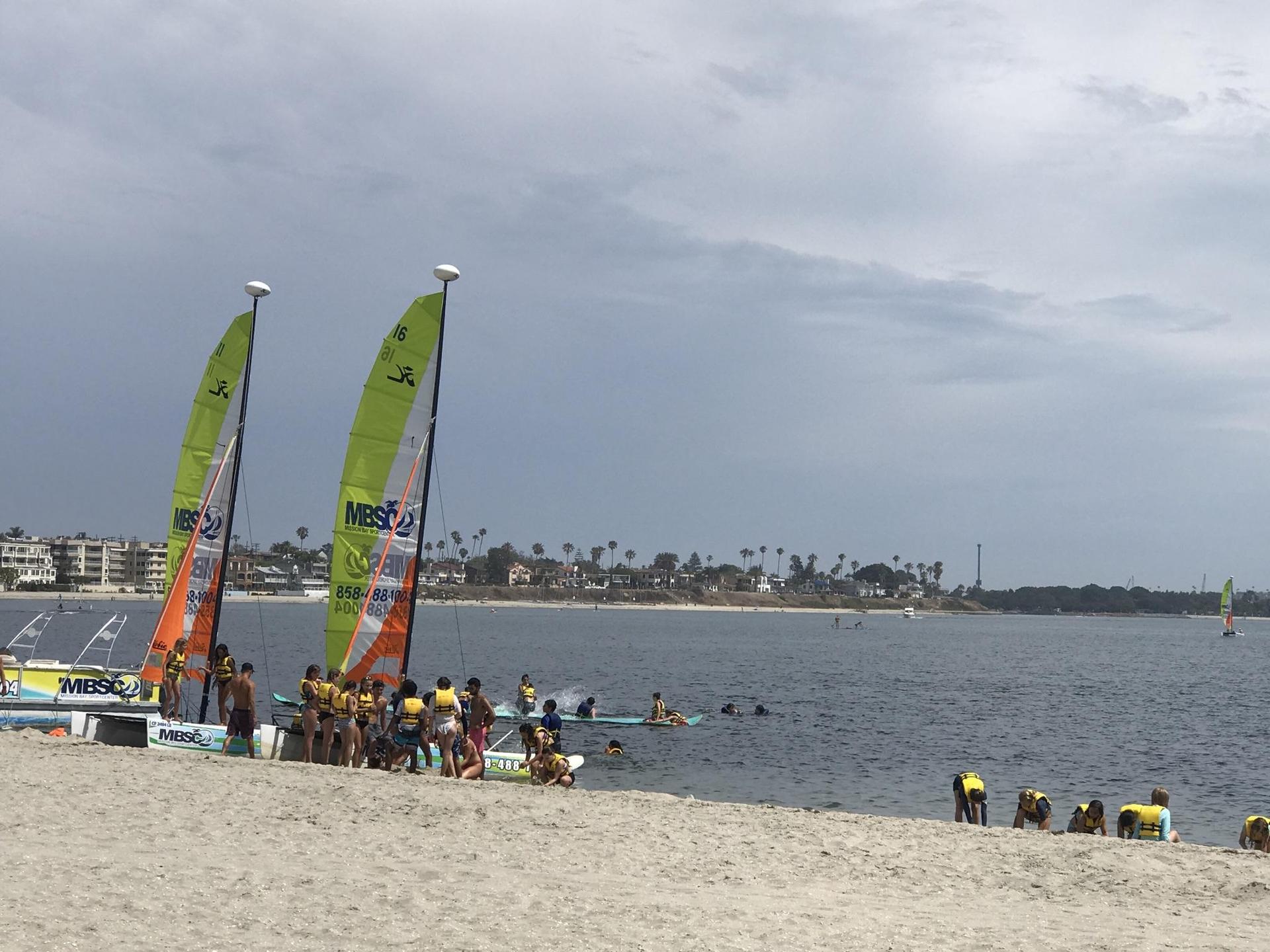 People gather around sailboats on a sandy beach next to calm water under a cloudy sky.
