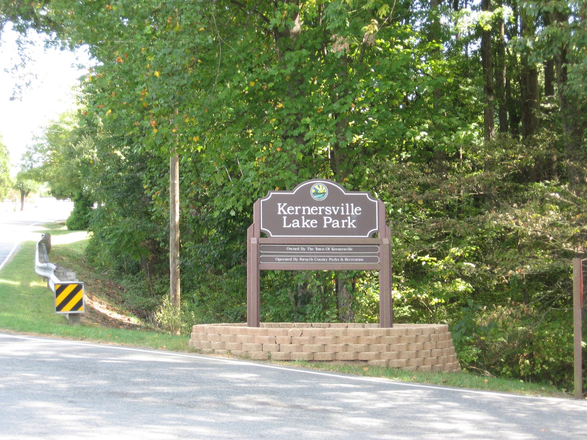 A wooden park entrance sign on a stone base, surrounded by green trees.