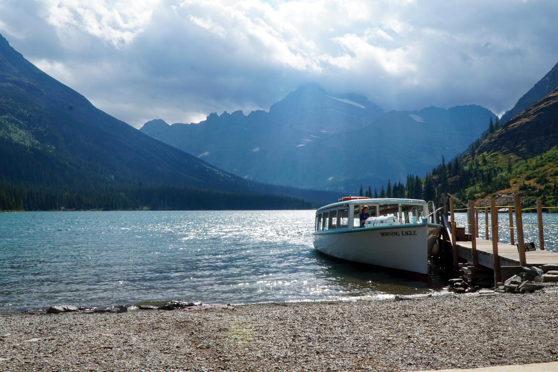 A tour boat docked on a lake, with dramatic mountains under a cloudy sky.