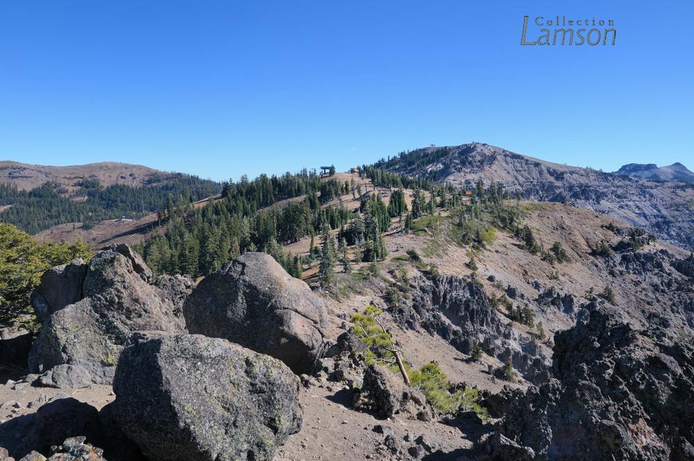 Sunny mountain landscape with large rocks, sparse trees, and distant peaks.