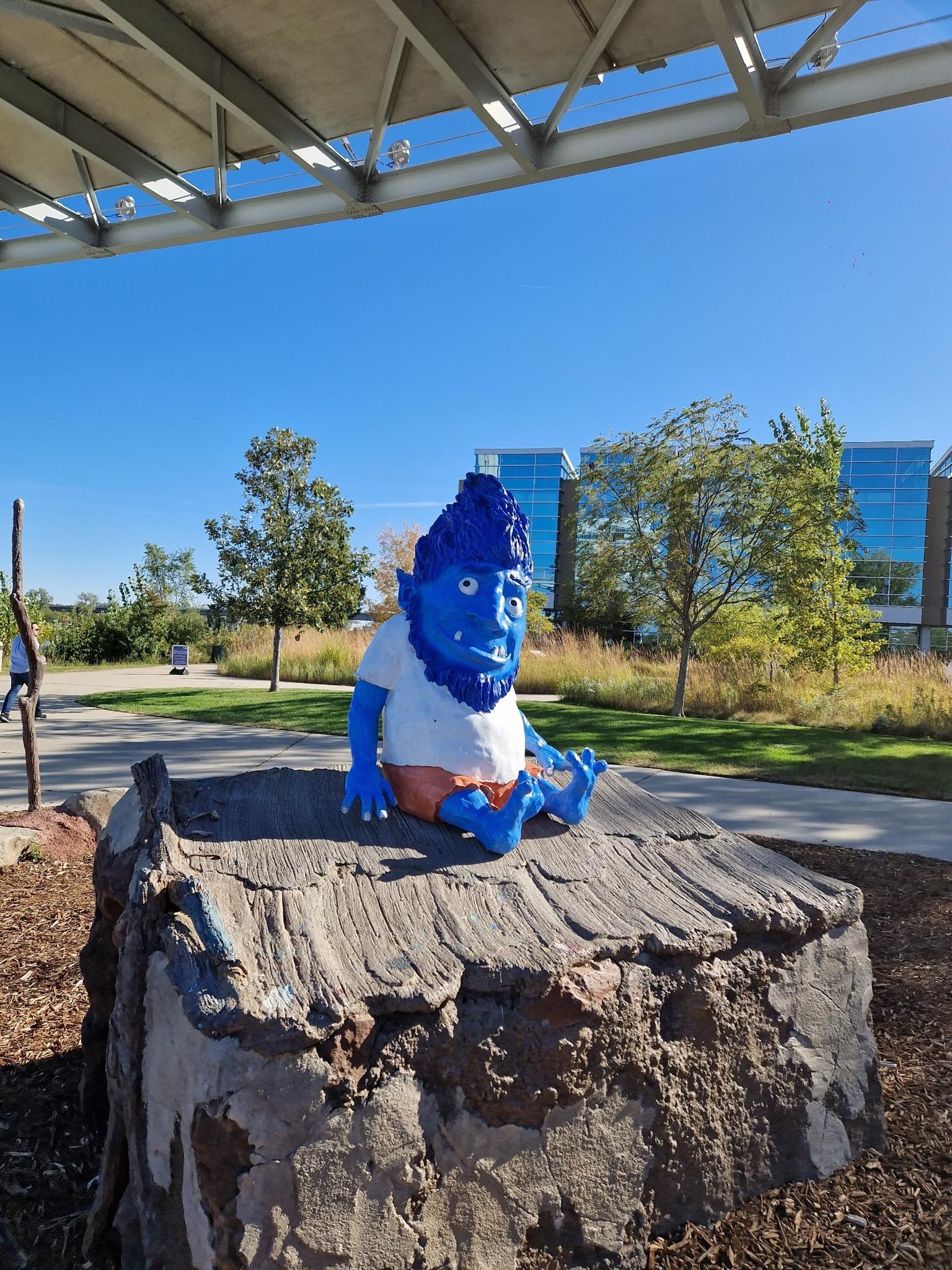 Blue gnome statue on a tree stump, with modern buildings in background.