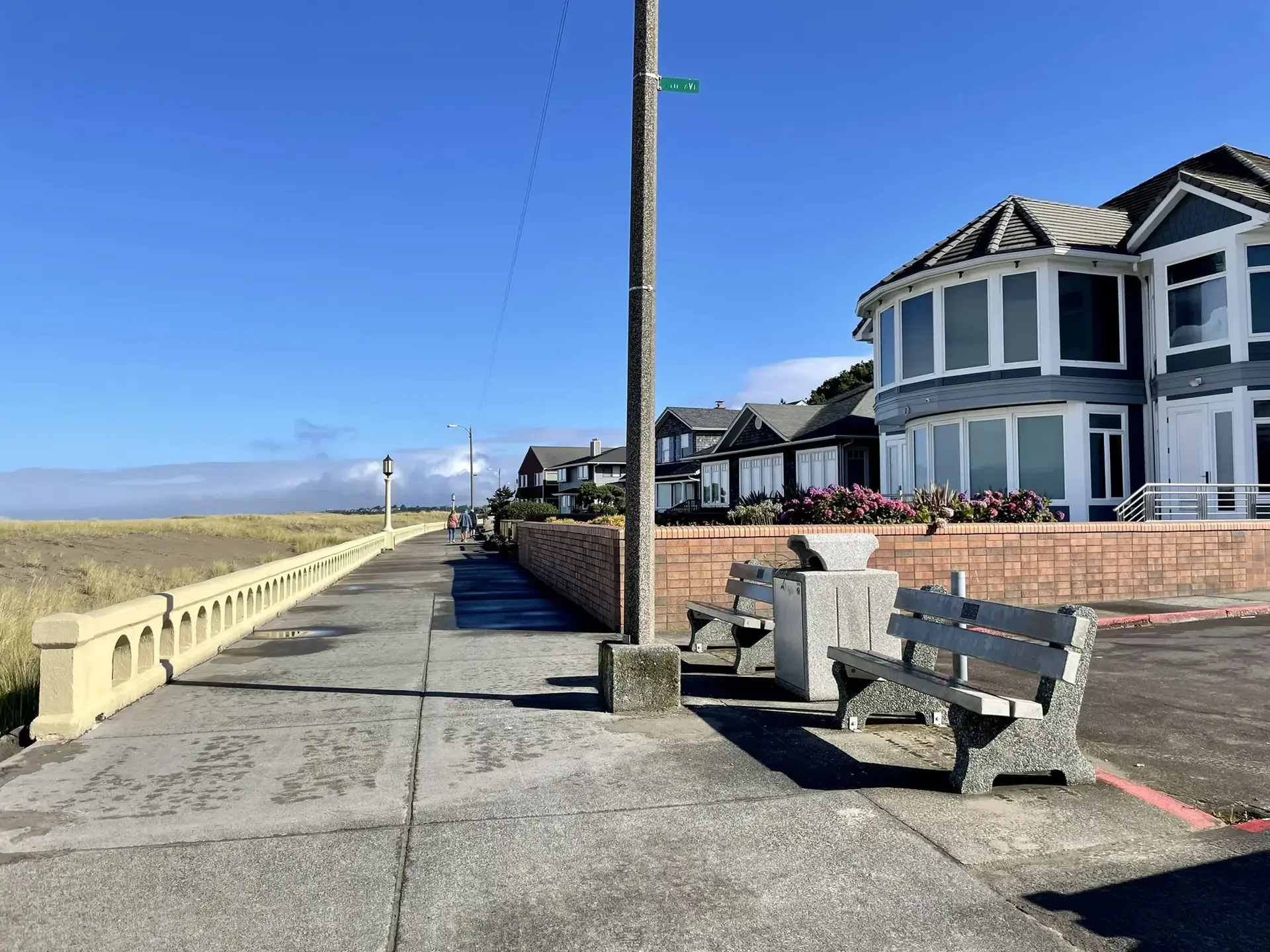Sunny boardwalk with benches, houses, and a distant grassy field.