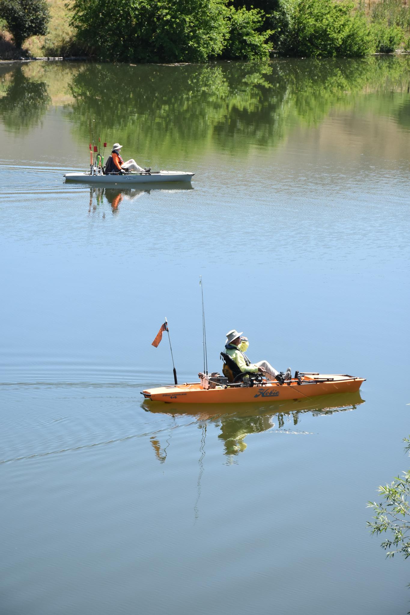 Two people kayak on calm water with reflections of green trees under bright sun.