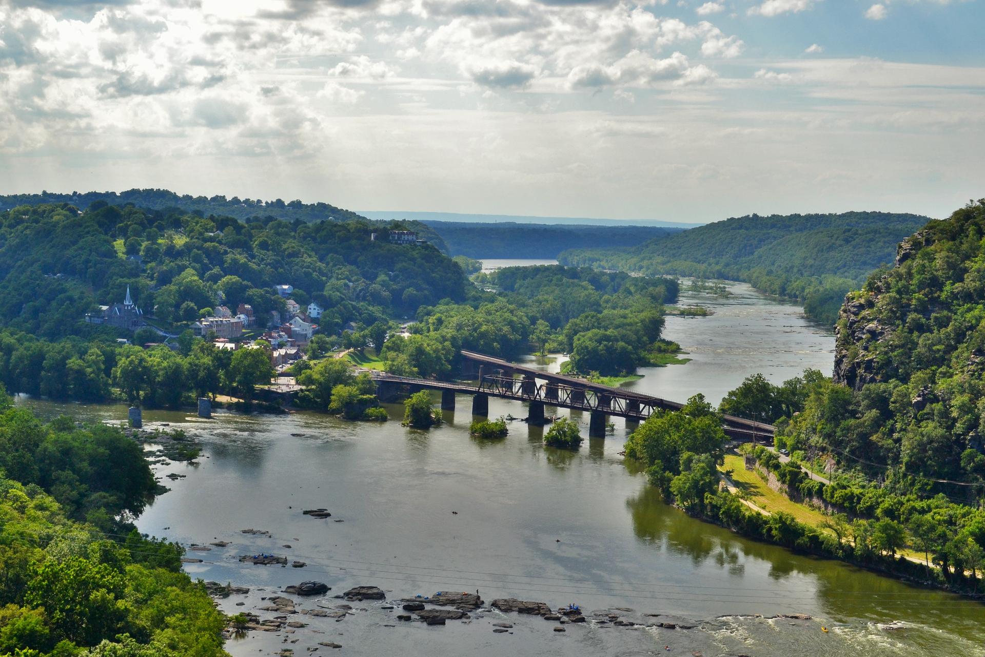 Expansive view of a wide river, a long bridge, and a town among green hills.