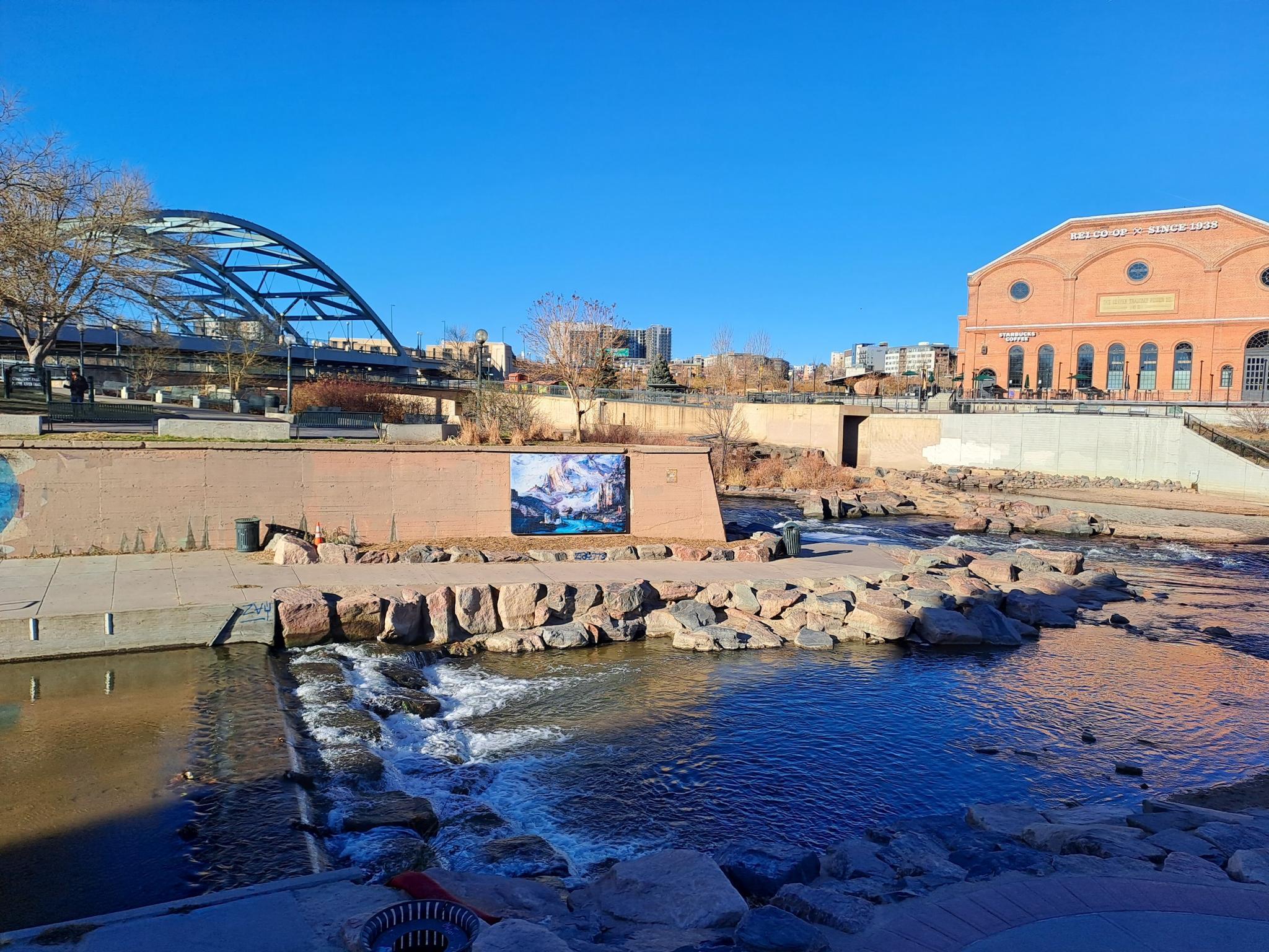 Urban river with a small waterfall, brick buildings, and a modern bridge.