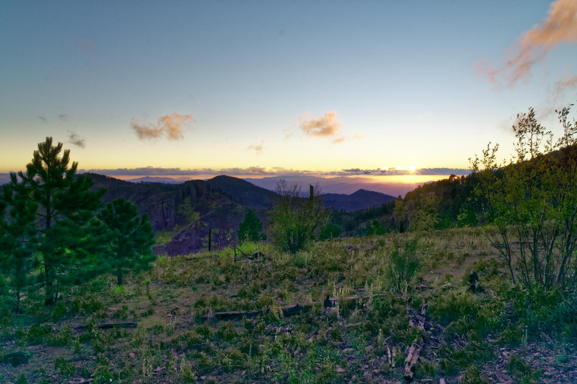 Mountain landscape at sunset with warm light on the horizon and scattered trees.