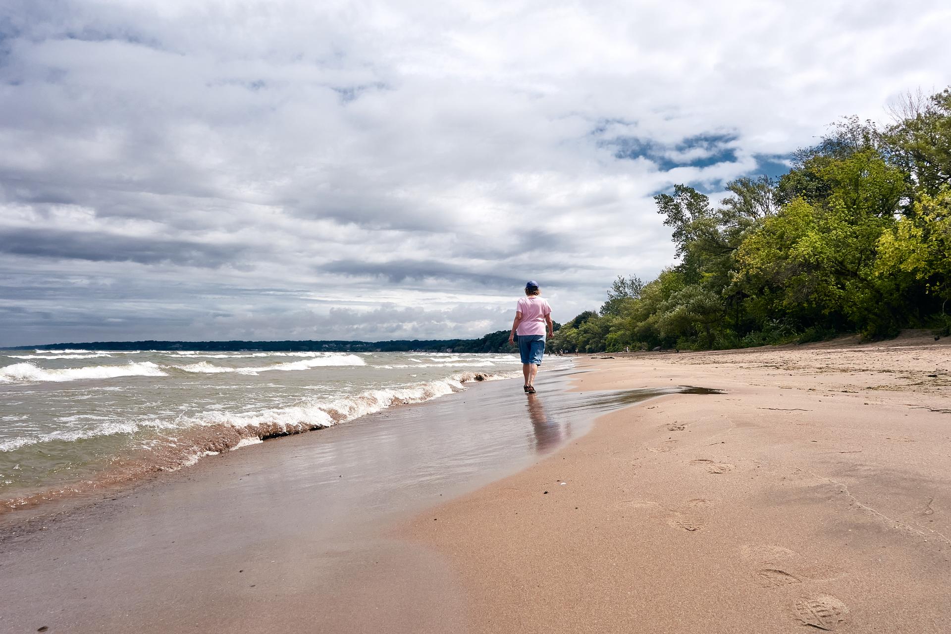 Person walks along a sandy beach with waves under a cloudy sky.