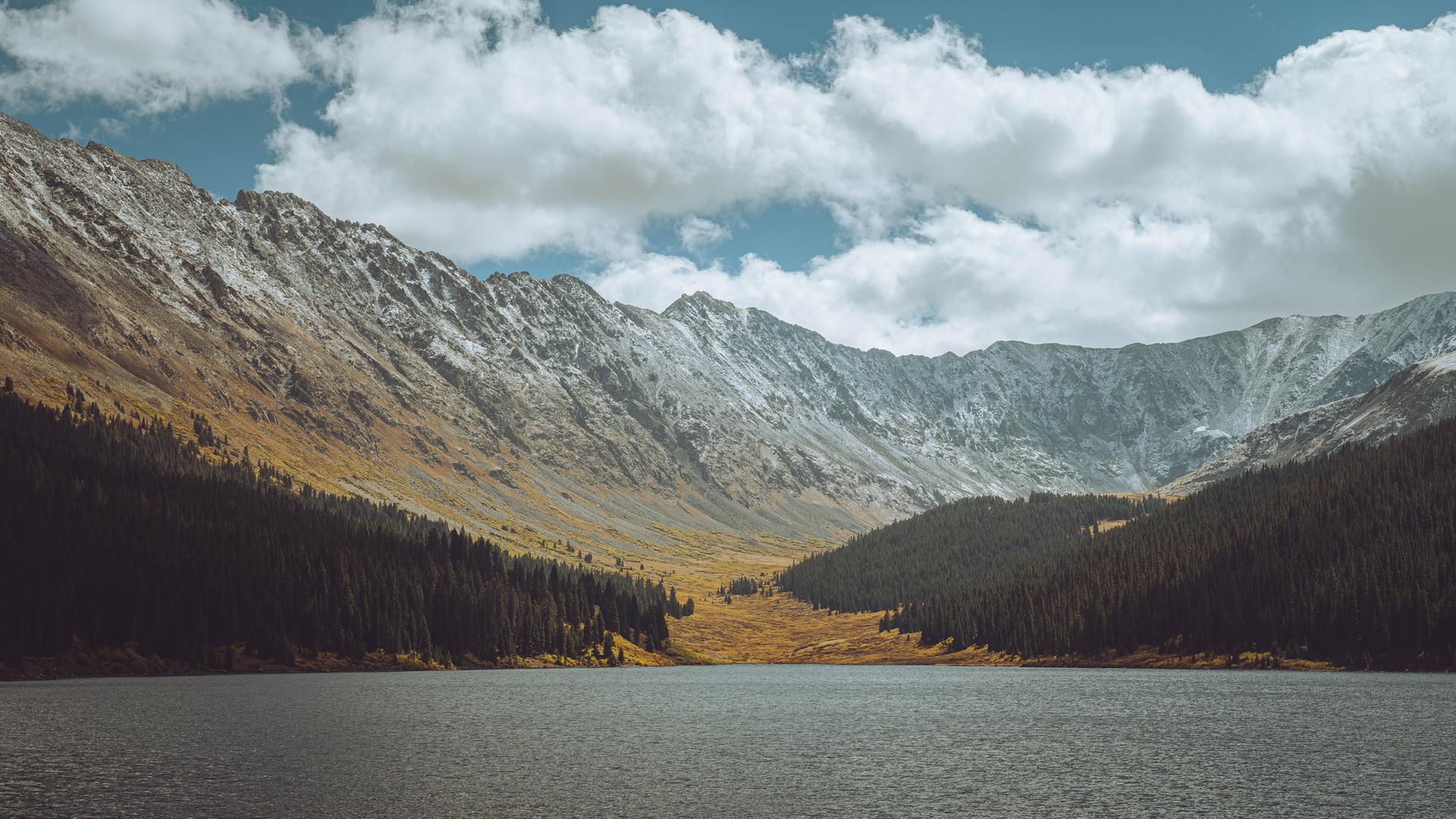 Serene mountain lake reflecting snow-dusted peaks and pine forests under clouds.