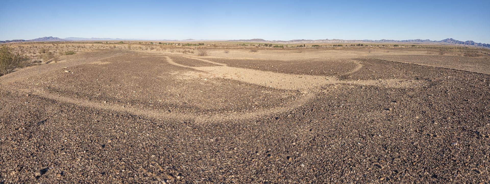 Vast, dry desert landscape with faint ground patterns under a clear blue sky.
