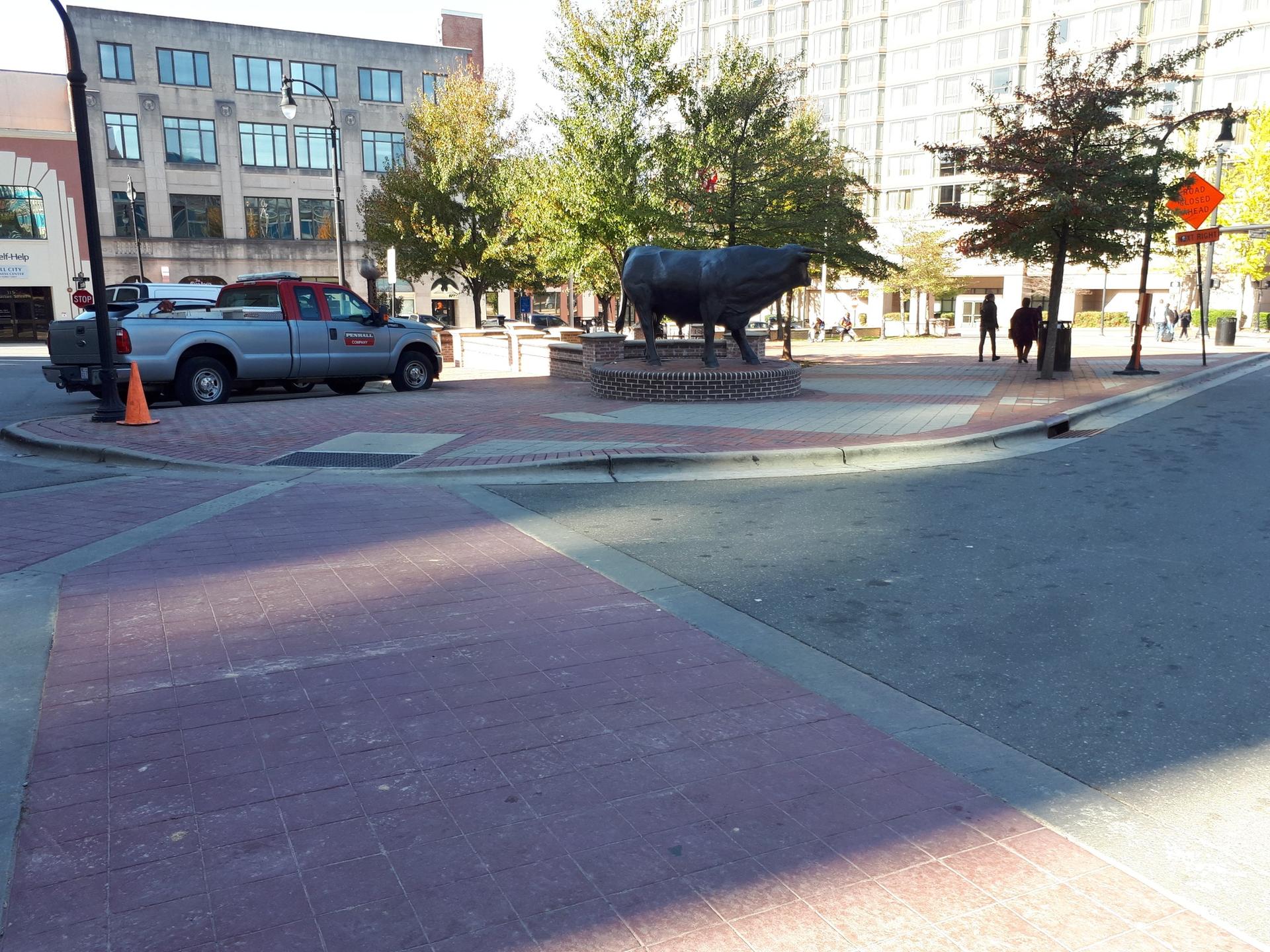 A large bull statue stands in a sunny urban square with buildings.