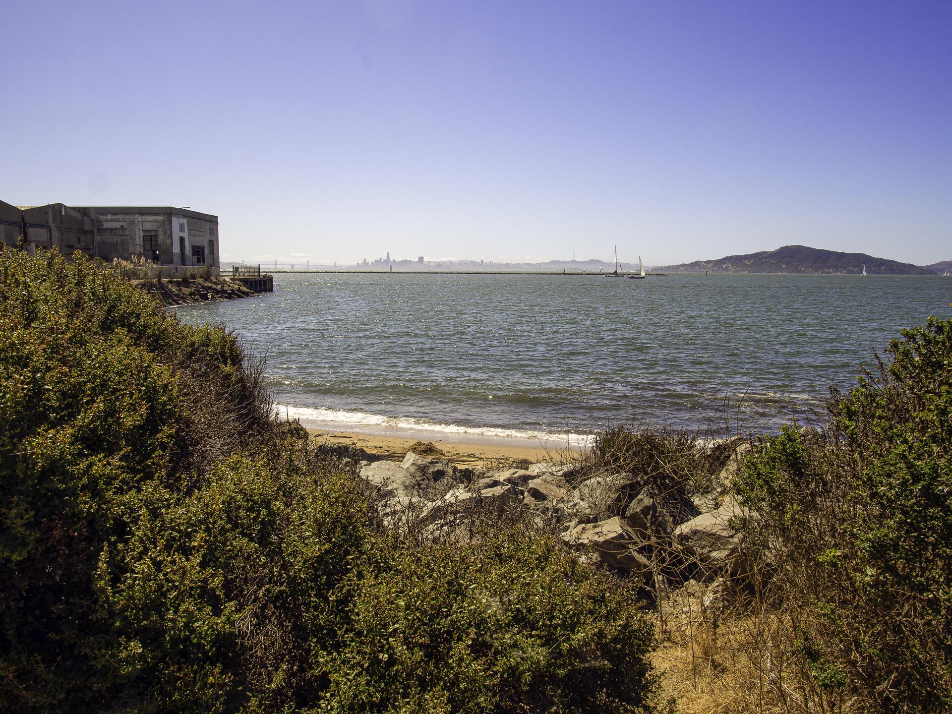 Sunny view of a bay with an old building, rocky beach, and distant land.