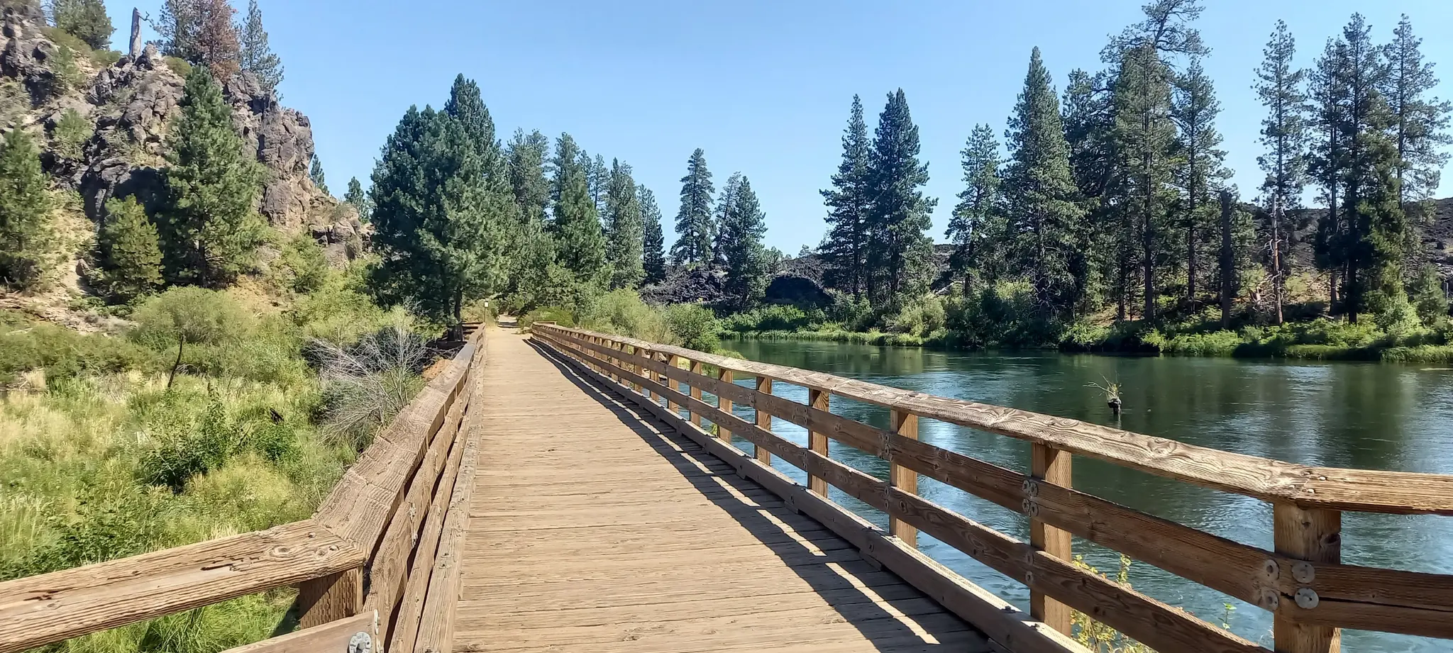 Wooden boardwalk alongside a calm river with trees under bright sunlight.