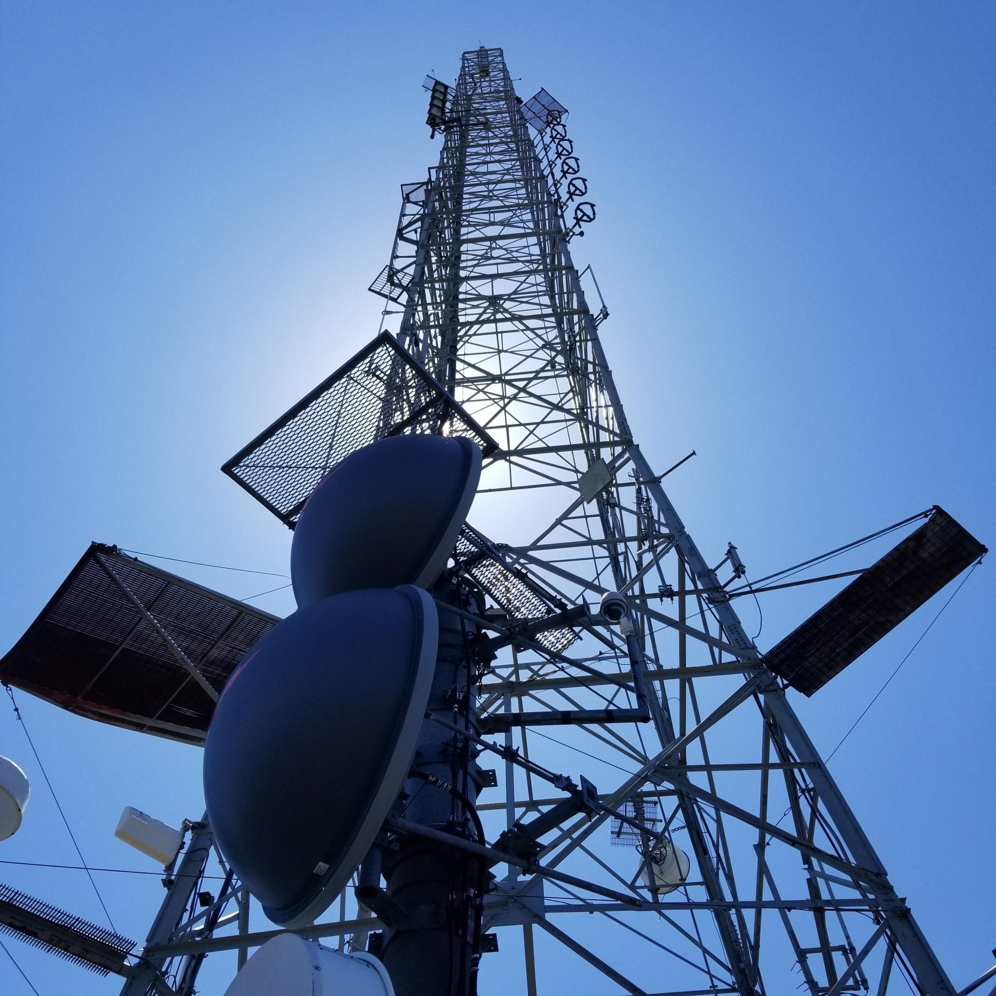 A tall metal communication tower against a bright blue sky with sun glare.