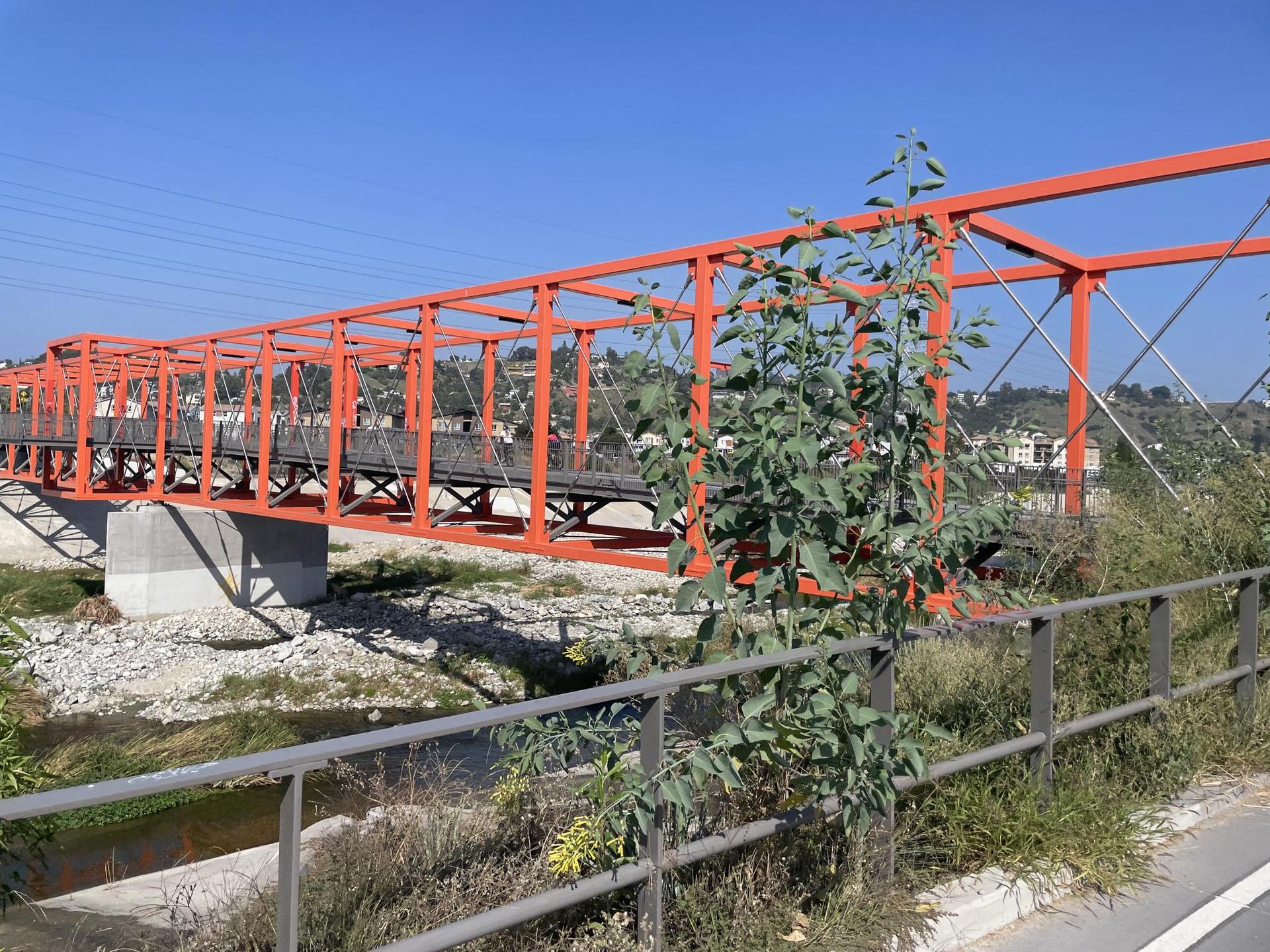 Bright orange truss bridge over a dry riverbed with distant hills, sunny.