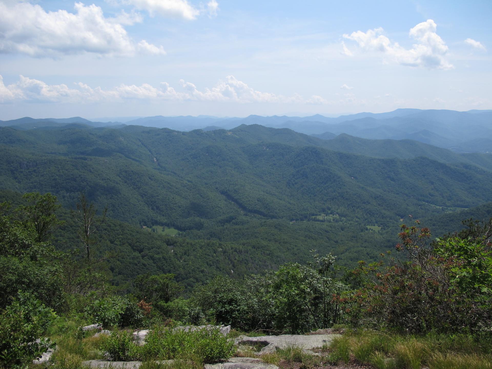 Expansive view of lush green forested mountains under a bright, cloudy sky.