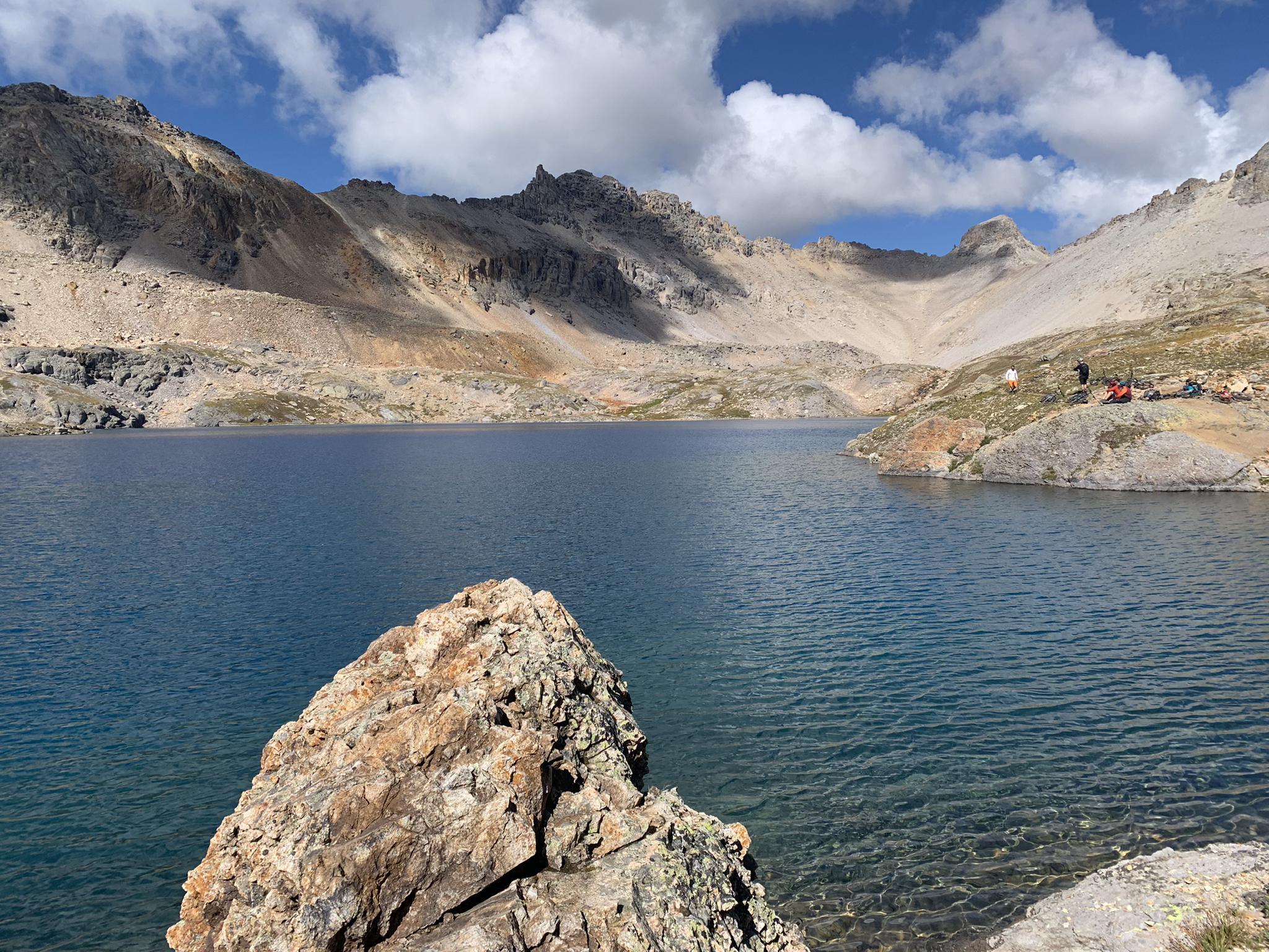Mountain lake with rocky shores, distant campers, and bright daylight under a cloudy sky.