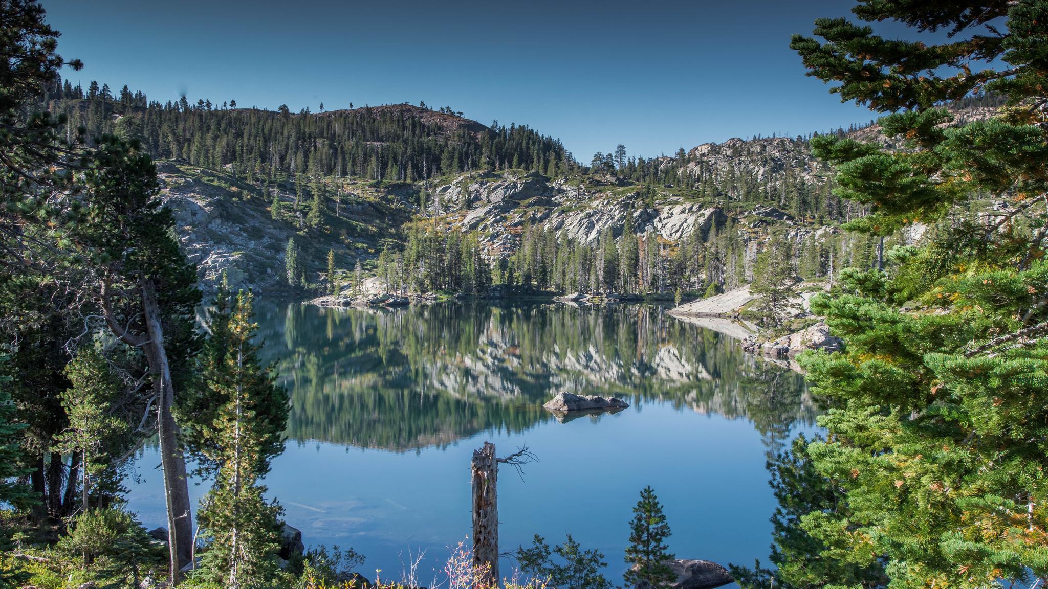 Serene lake reflecting pine trees and rocky hills, framed by lush green trees.