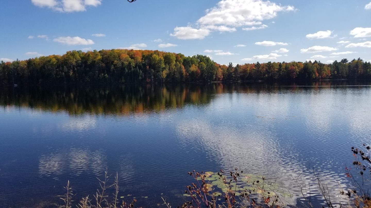 Wide lake with colorful autumn trees reflecting on calm water under blue sky. - Bruno's Run Trail