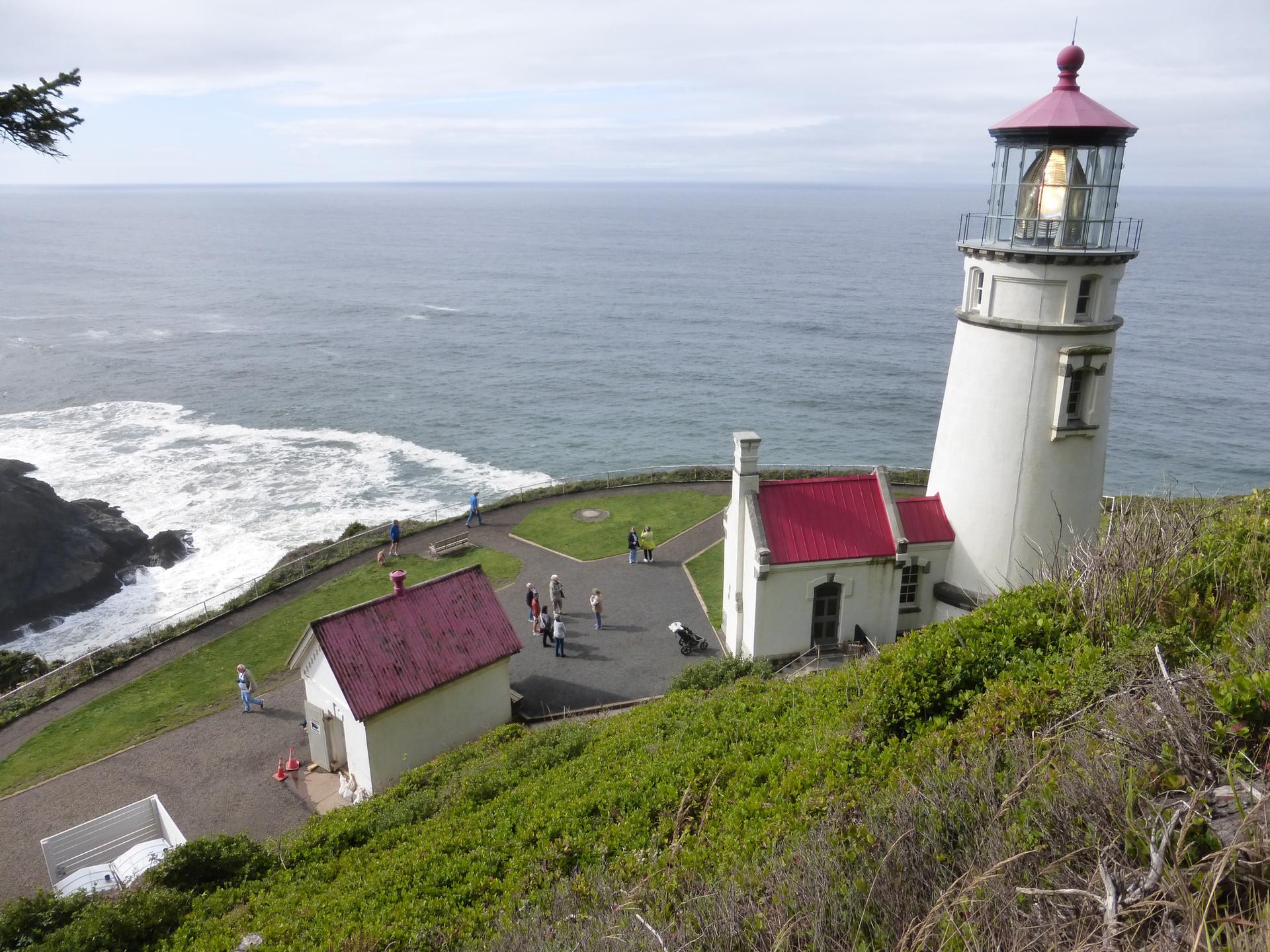 White lighthouse and buildings on a green cliff overlooking the ocean, with people.