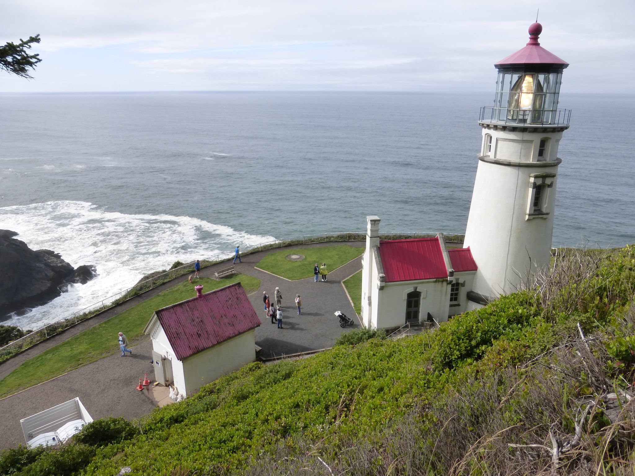 White lighthouse and buildings on a green cliff overlooking the ocean, with people. - Heceta Lighthouse Trail