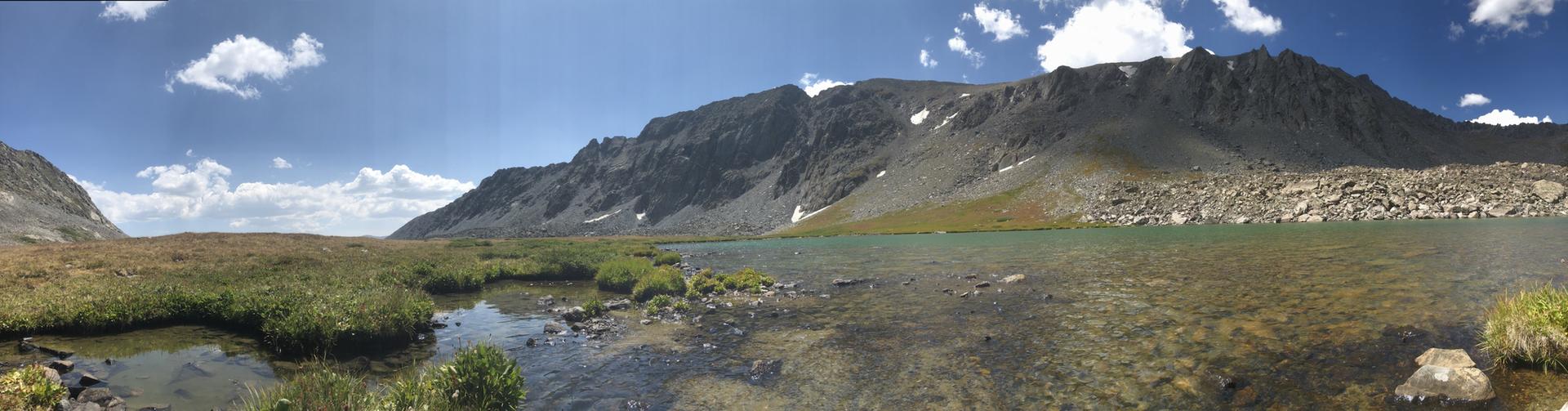 Panoramic view of a clear lake and vast rocky mountains under blue sky.
