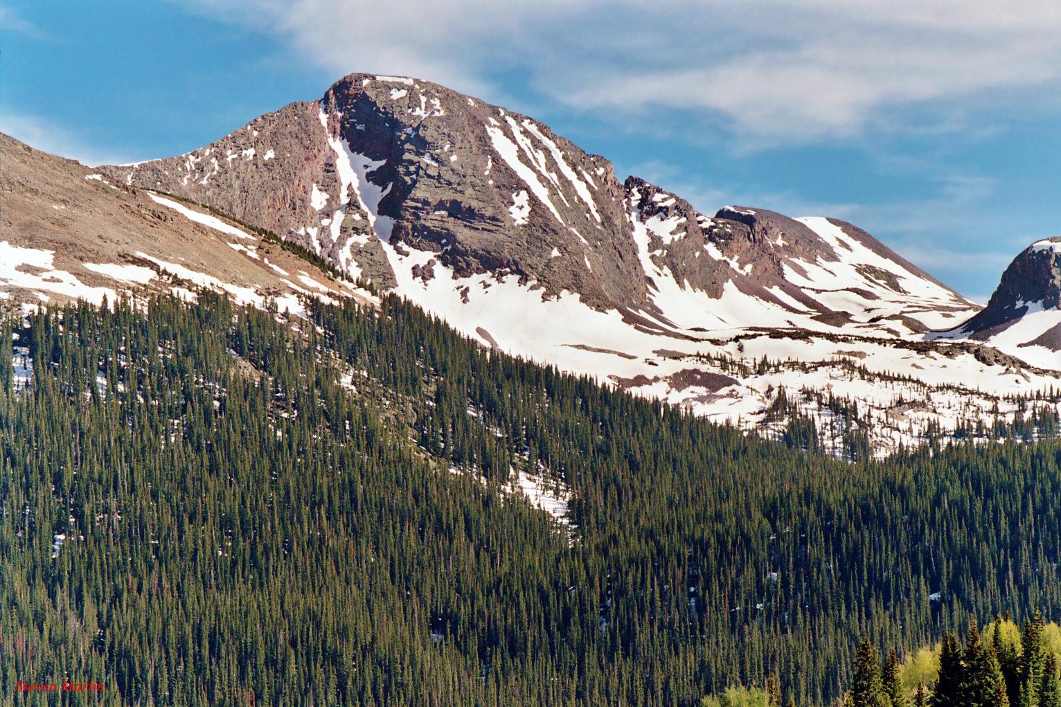 Rugged mountain with snow patches above a dense evergreen forest on a clear day.