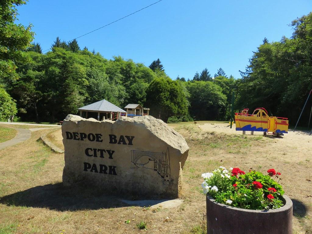 Sunny park scene with a stone sign, playground, and lush green trees.