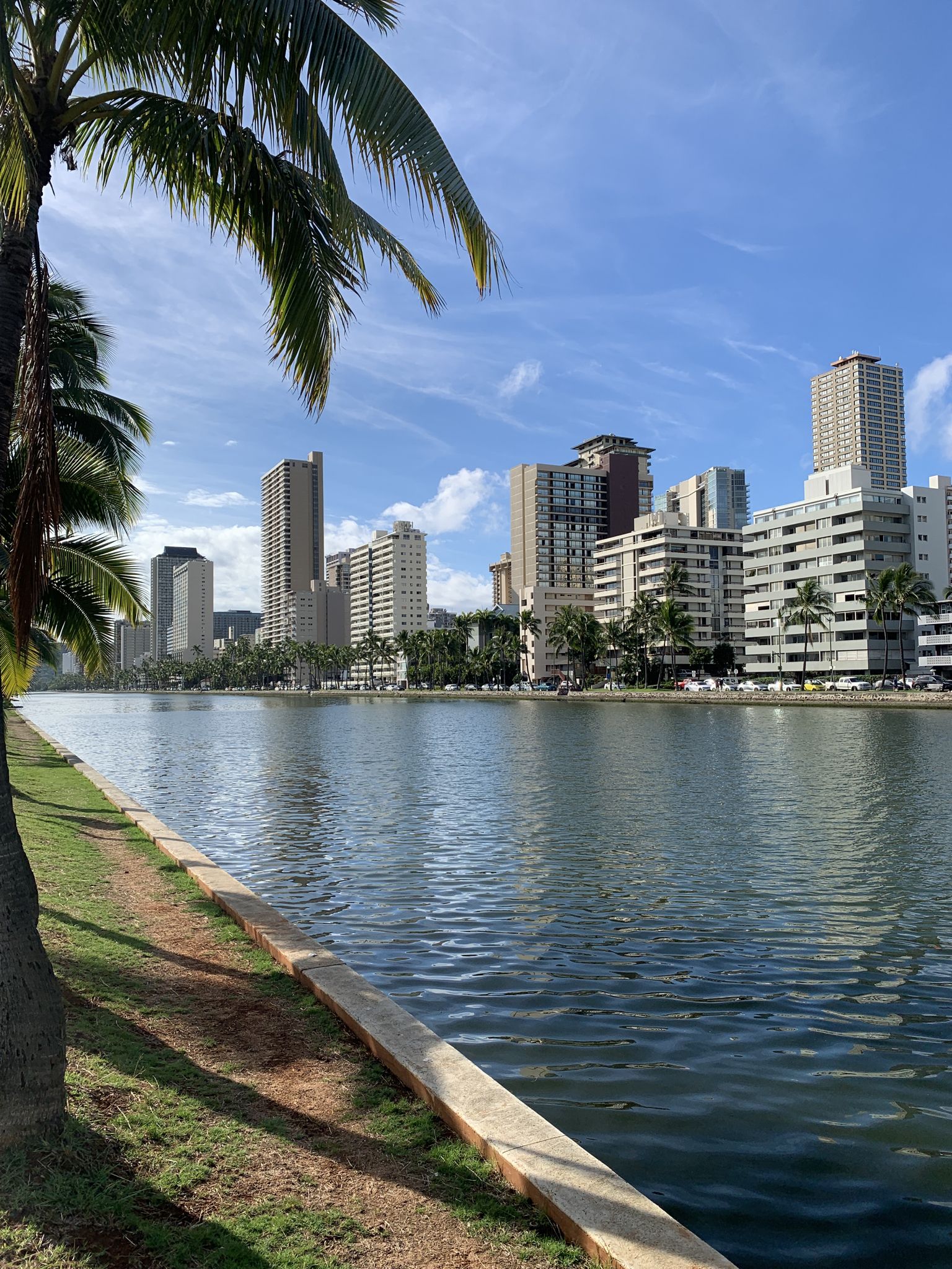 City skyline reflected in calm water under a bright blue sky. - Ala Wai Park Trail