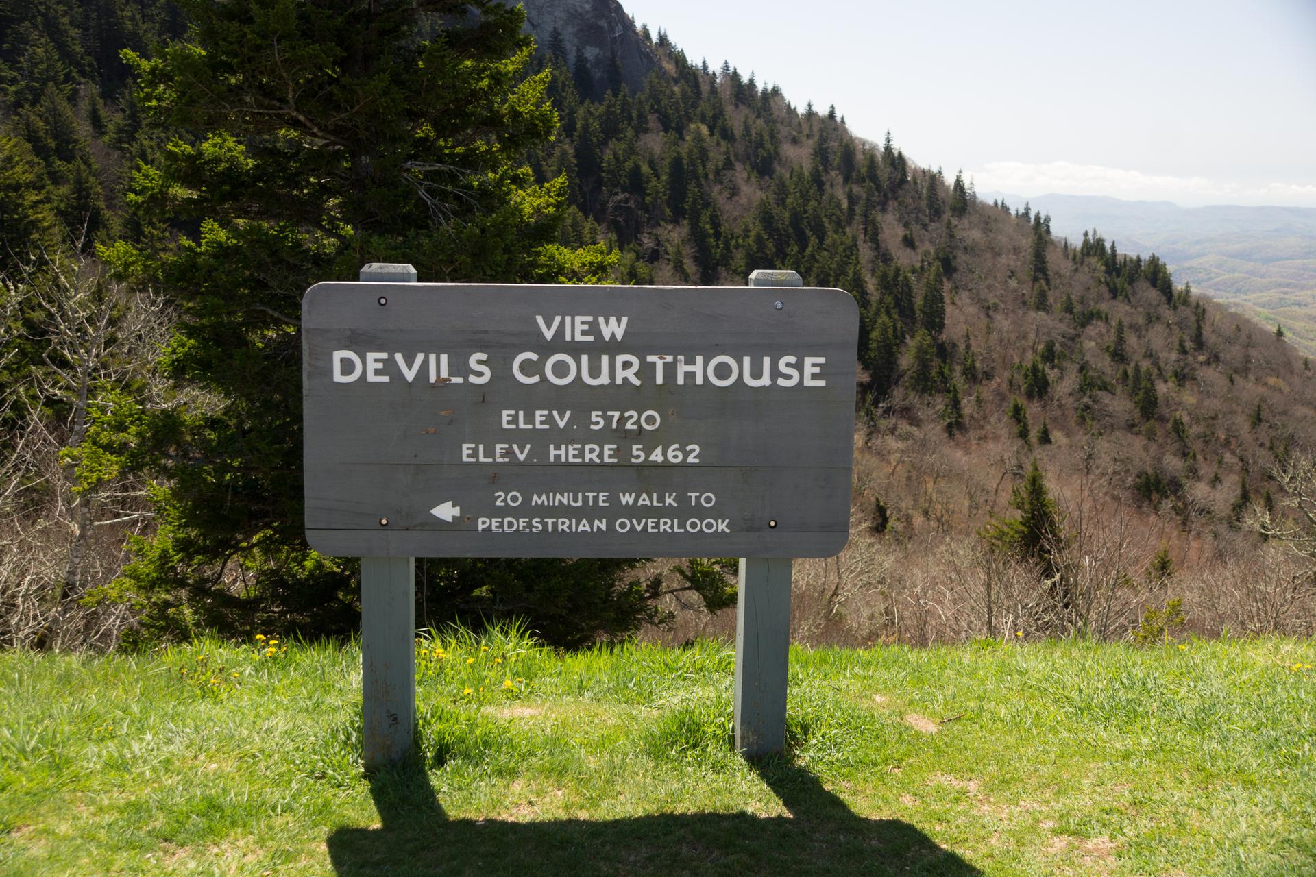 A wooden sign stands on a grassy hill overlooking a forested mountain.