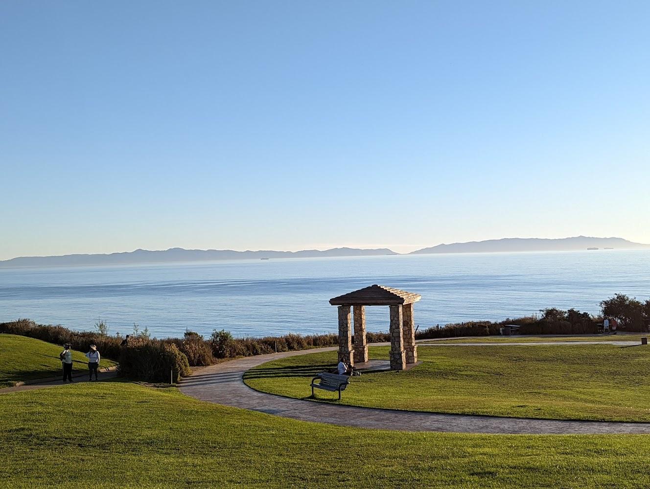 Grassy park with a stone gazebo, ocean view, and people walking.