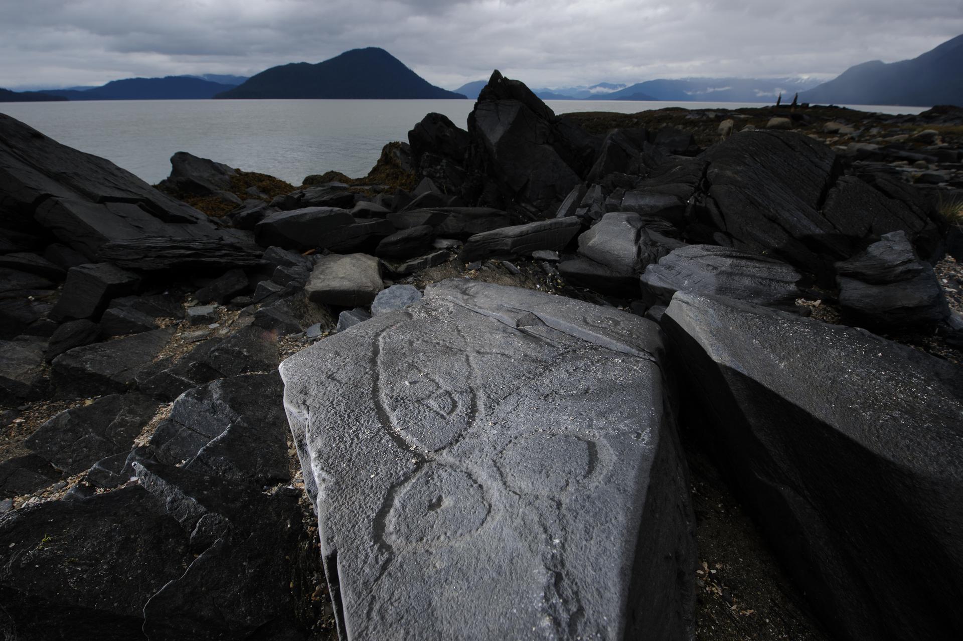 Ancient carvings on dark rocks by water, distant mountains, and small figures.